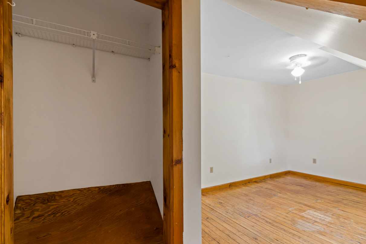Interior view of a closet and bedroom. Closet has wooden trim, a shelf and empty space. Bedroom has wood floor, white walls and a ceiling light.