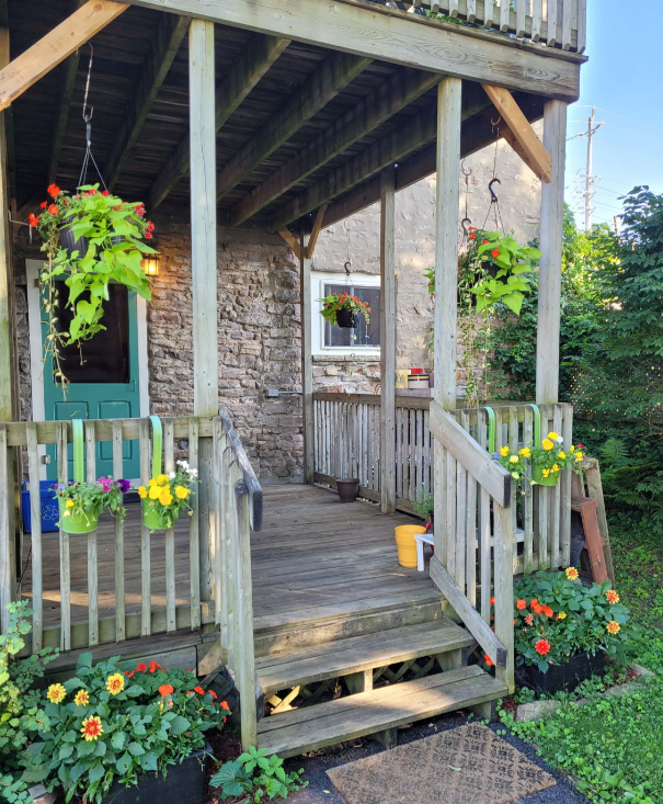 Wooden porch with hanging flowers, steps, and a turquoise door against a stone wall.