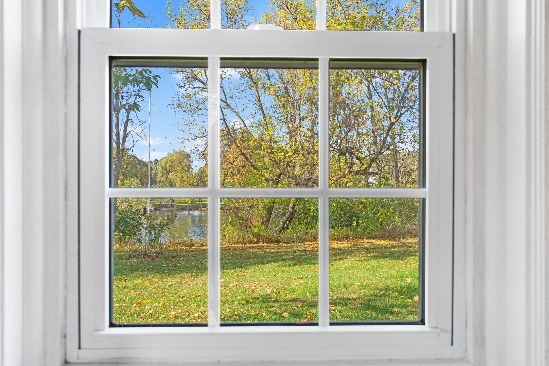 White window looking out onto a grassy yard with trees and a body of water in the distance, blue sky.