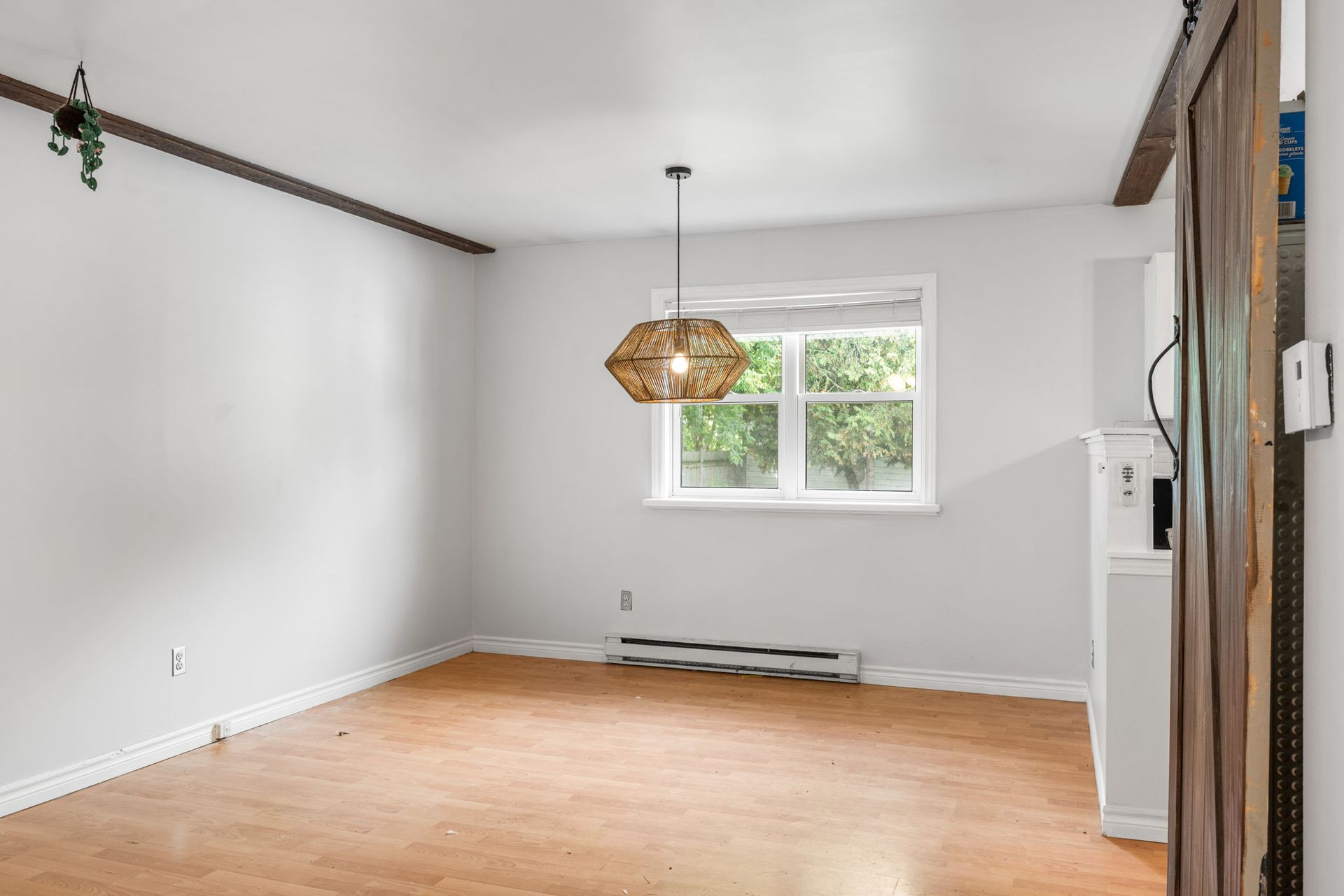 Empty room with wood flooring, white walls, and a window with greenery. A pendant light hangs from the ceiling.