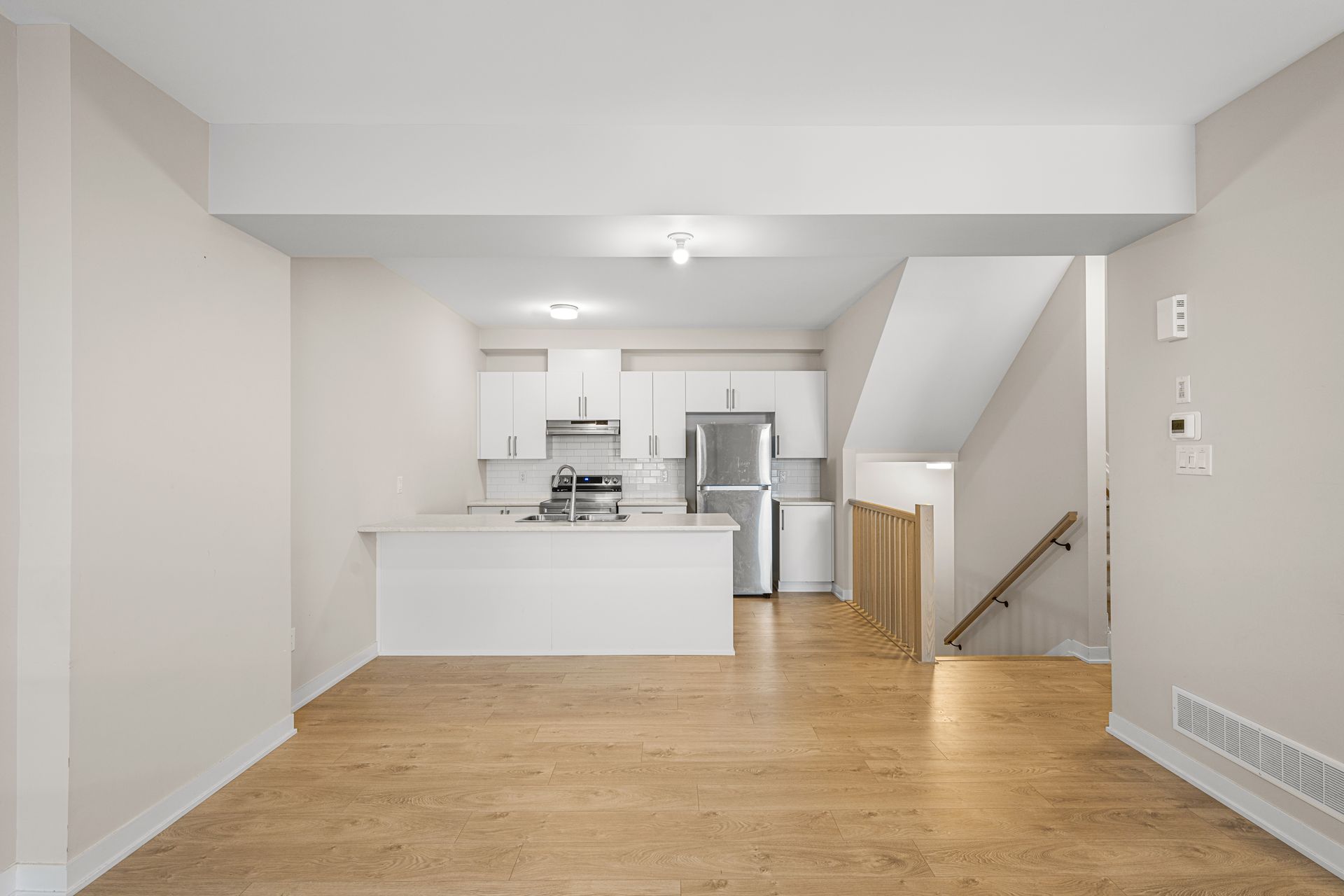 An empty living room with hardwood floors and a kitchen in the background.