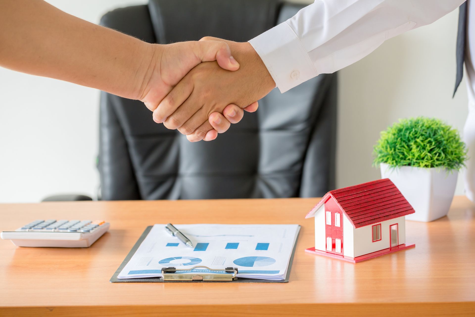 Two people shaking hands over a desk with paperwork, a toy house, and a calculator.