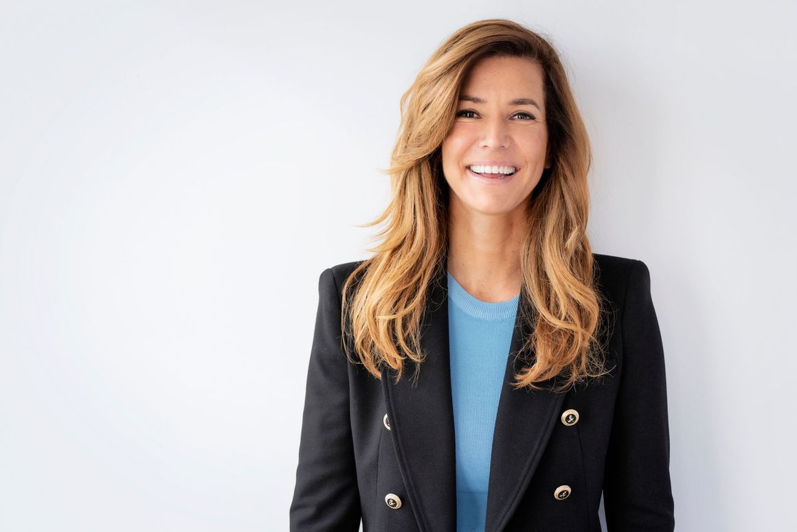 A woman in a black jacket and blue sweater is smiling while standing in front of a white wall.