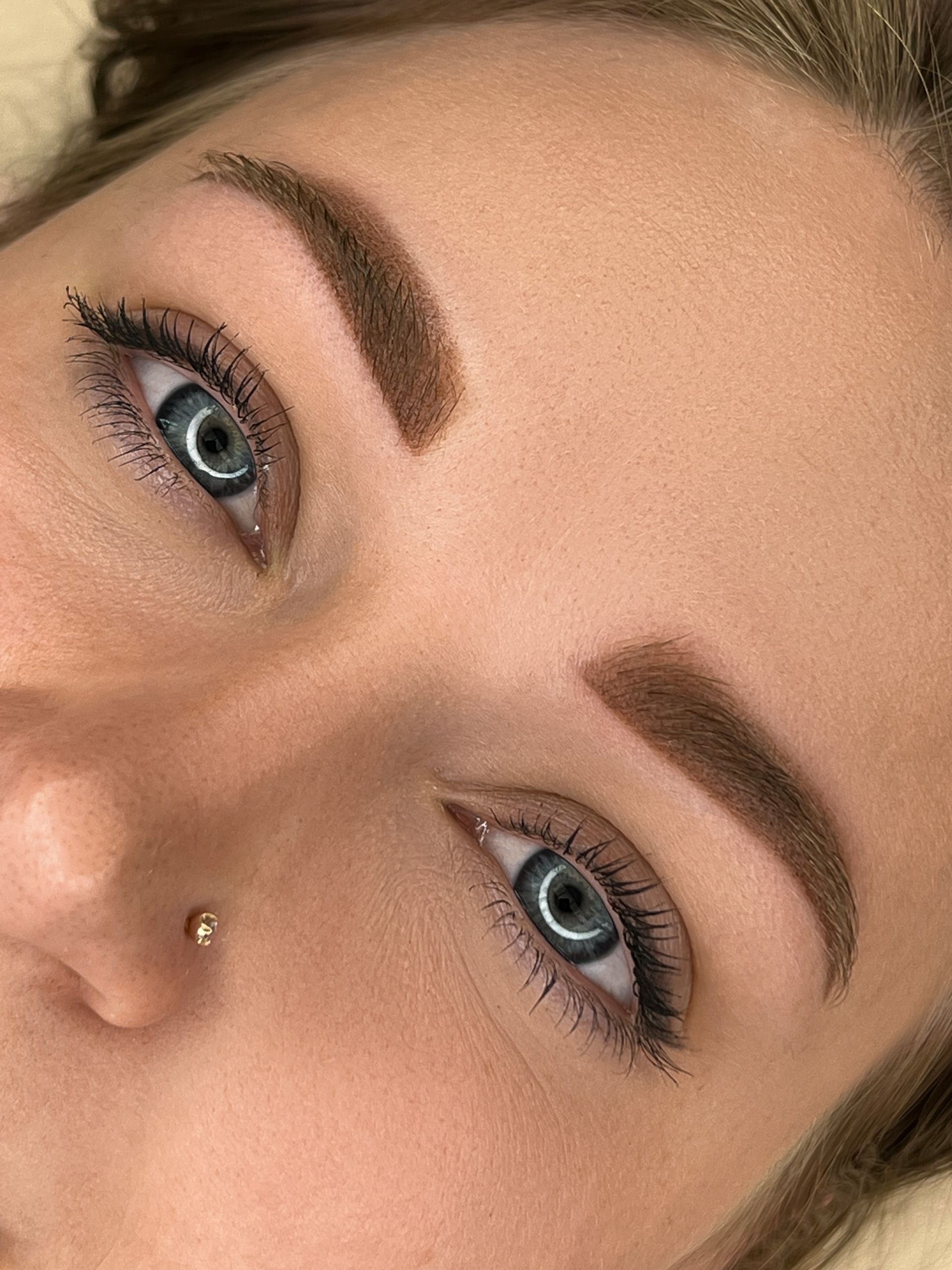 Nahaufnahme eines Gesichts mit blauen Augen, definierten Augenbrauen und einem kleinen Nasenpiercing.