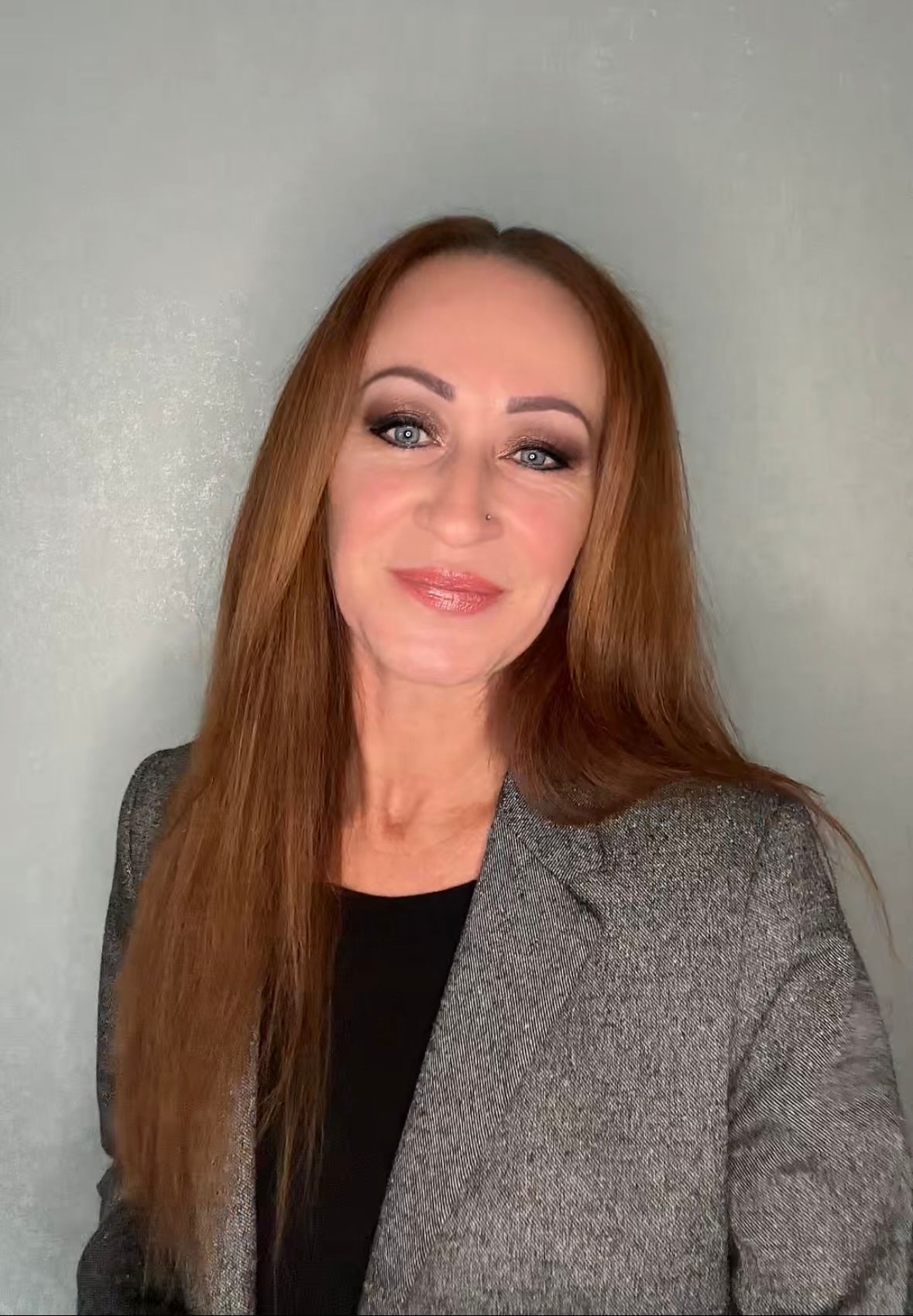 Person mit langen braunen Haaren, die einen grauen Blazer und ein schwarzes Oberteil trägt, vor hellgrauem Hintergrund.