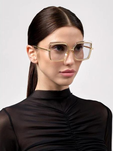 Woman wearing square, gold-rimmed glasses, dark hair pulled back, and a black mesh top.