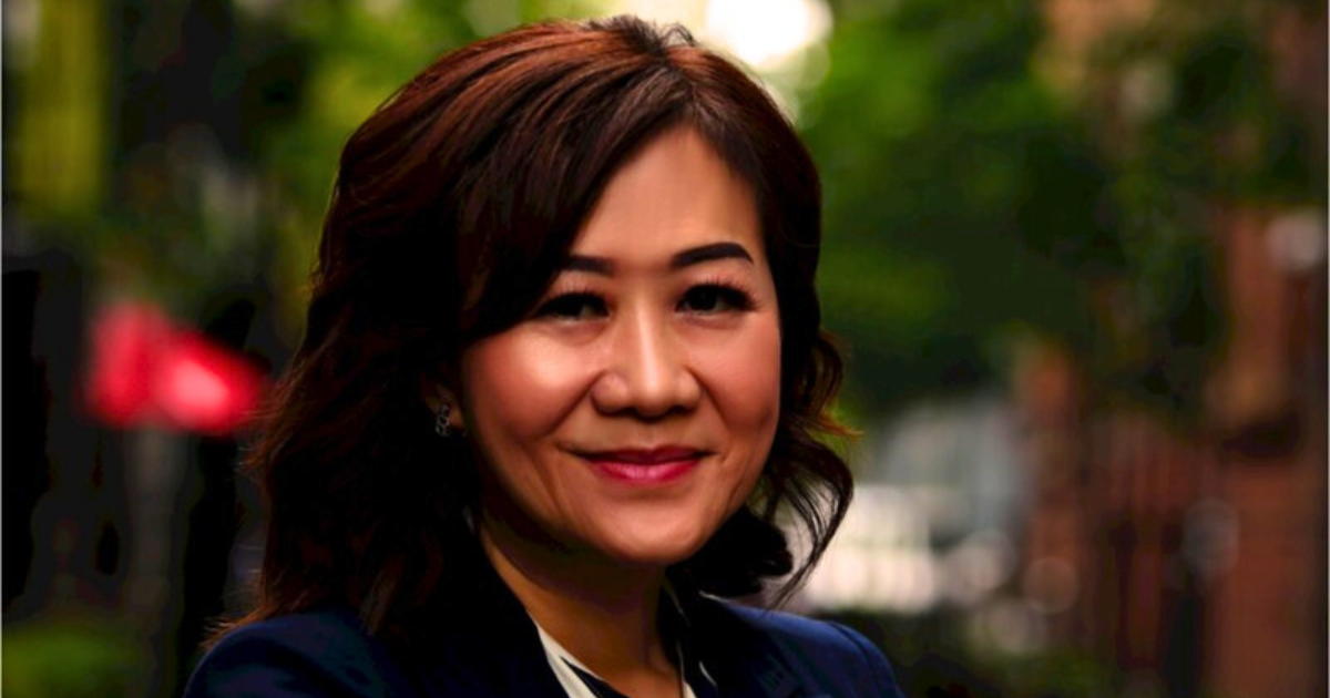 Woman in navy blazer smiles, arms crossed, in a city alleyway with trees.