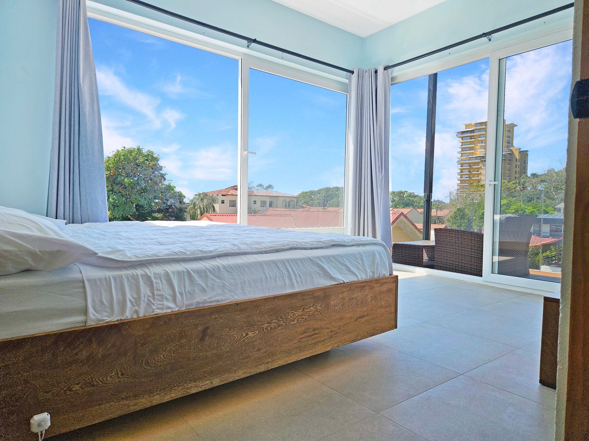 Bedroom with large windows, bed, light blue walls, and a sunny outdoor view.