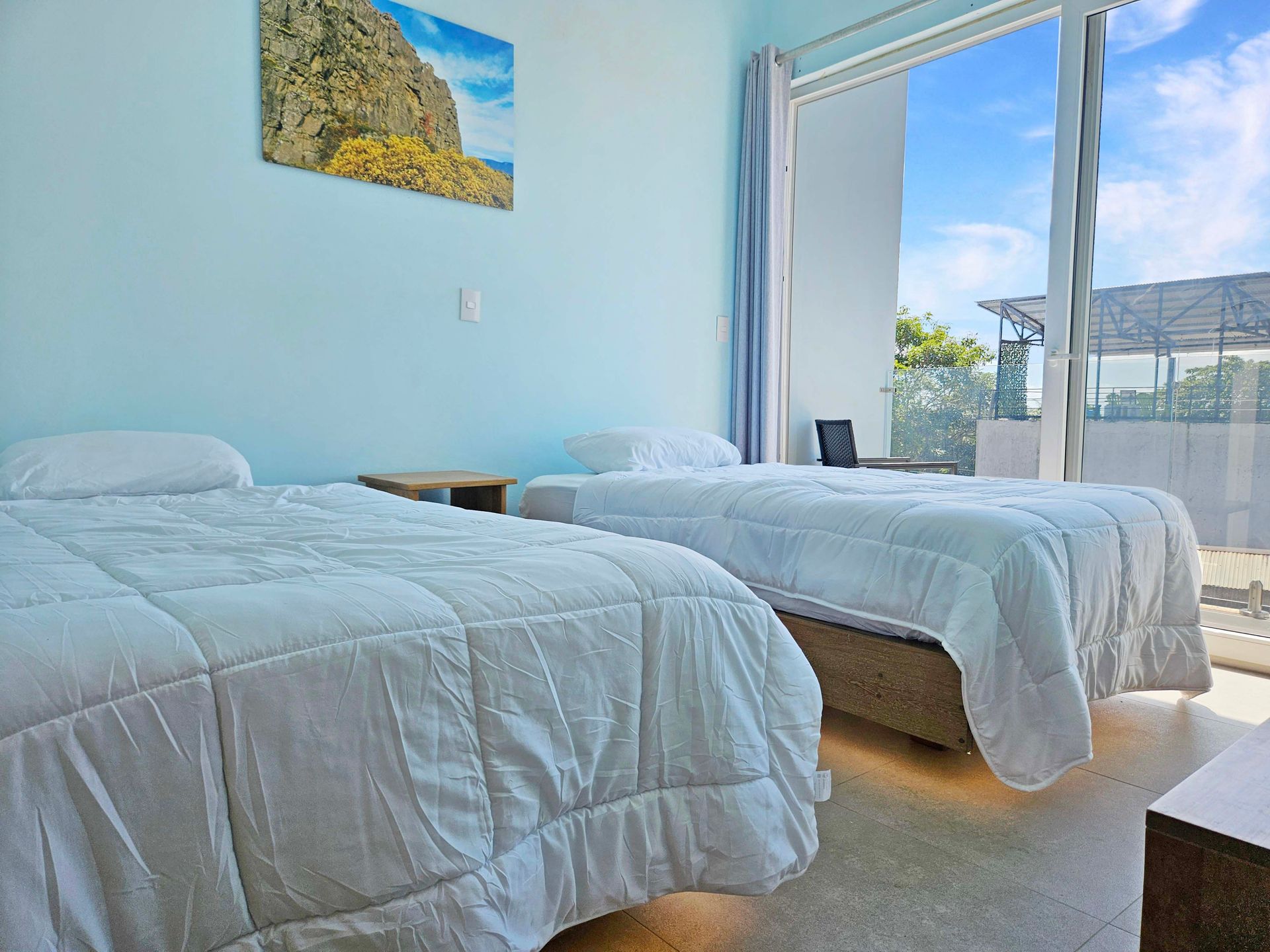 Two beds in a light blue bedroom with a balcony overlooking a blue sky and sea view.