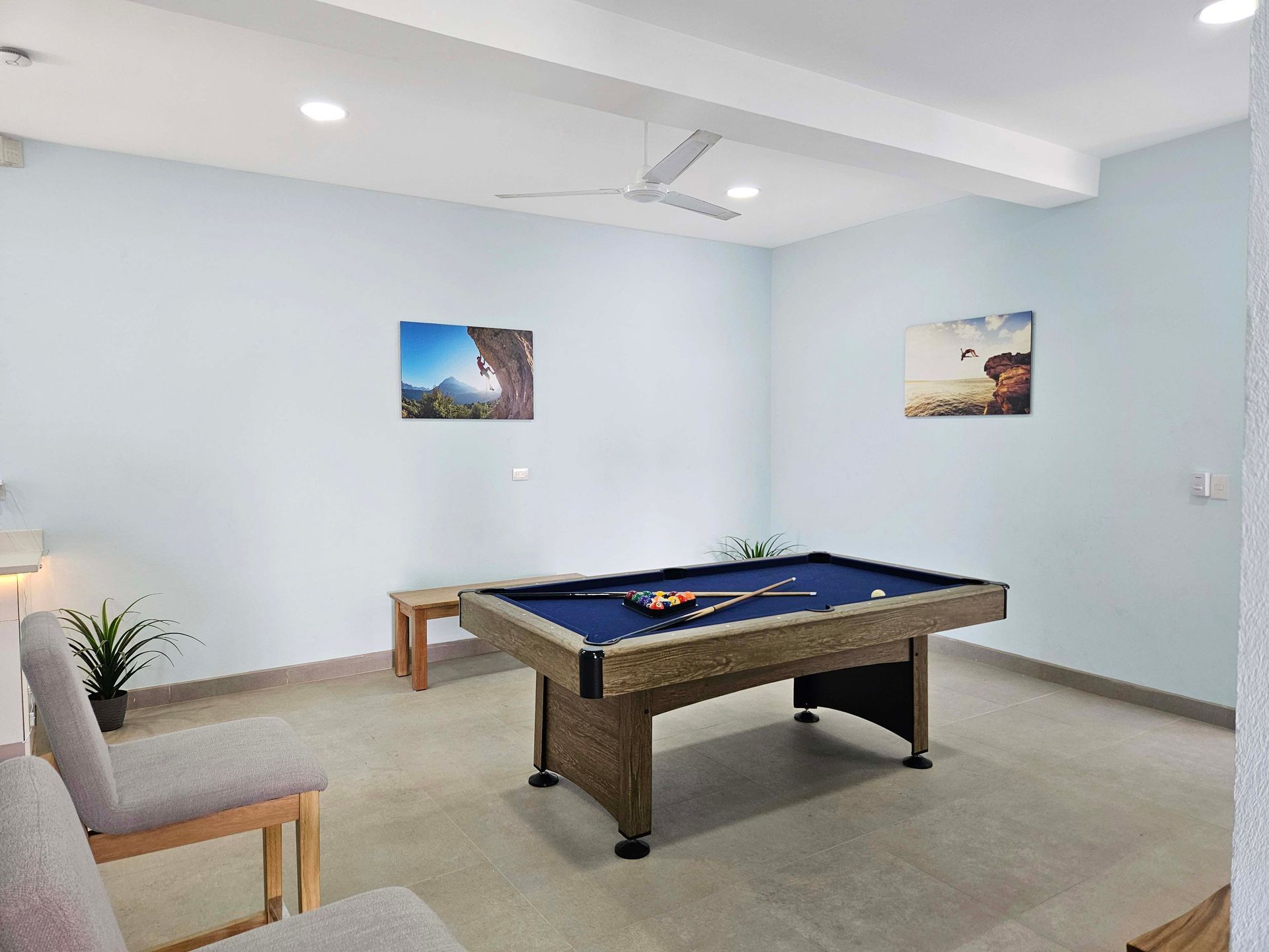 Pool table in a room with light blue walls, two paintings, chairs, and a small table.