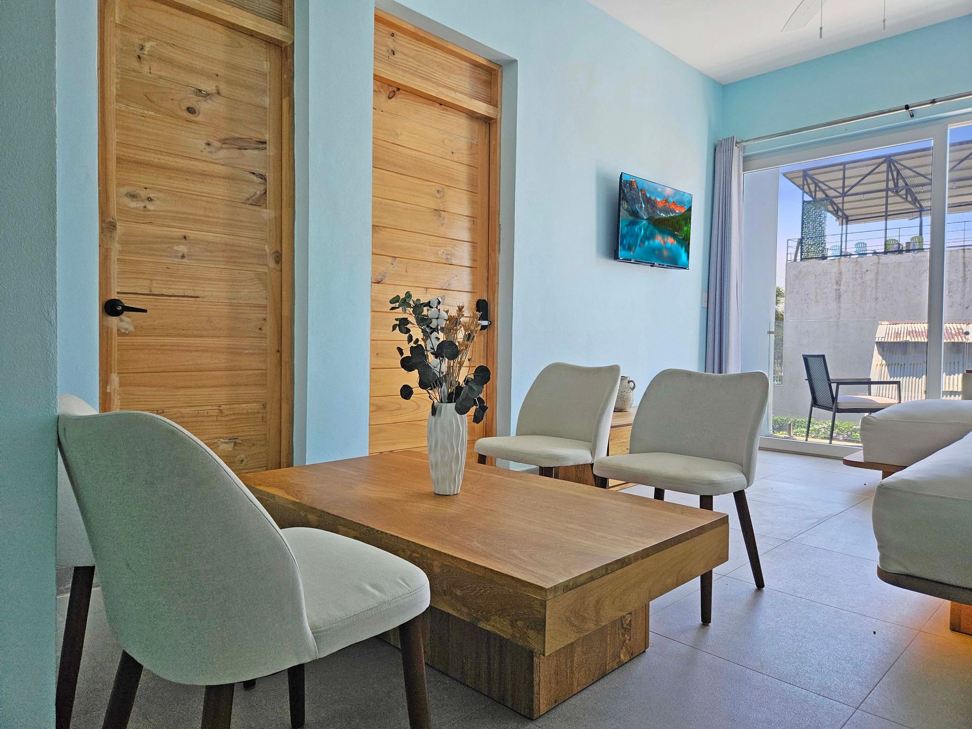 Living room with light blue walls, wooden doors, a coffee table, white chairs, and a TV.