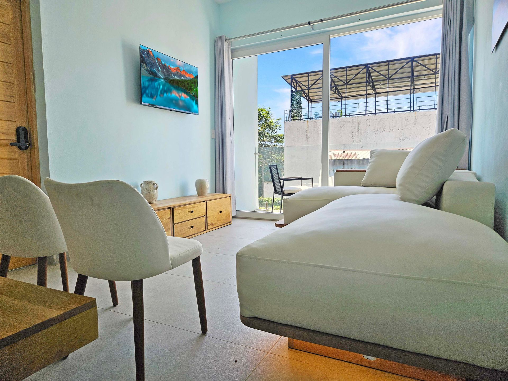 Bright living room with white sofa, chairs, wooden cabinet, and sliding glass doors leading to a balcony.