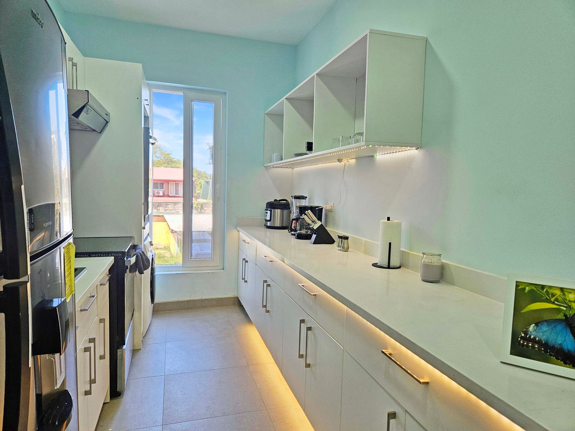 Long, narrow kitchen with white cabinets, countertop, and appliances. Blue walls, window, and overhead shelves.