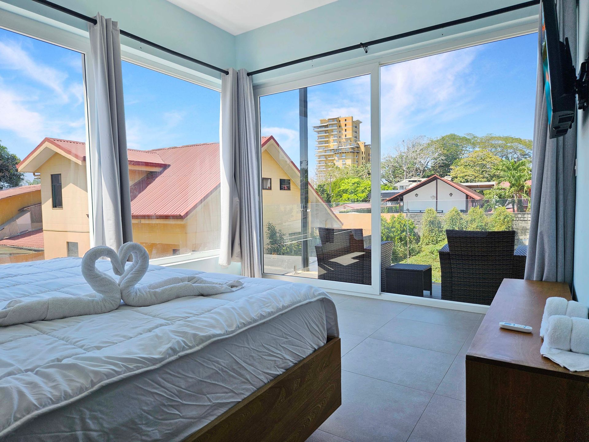 Bedroom with large windows, bed with swan towel art, and a view of buildings and trees.