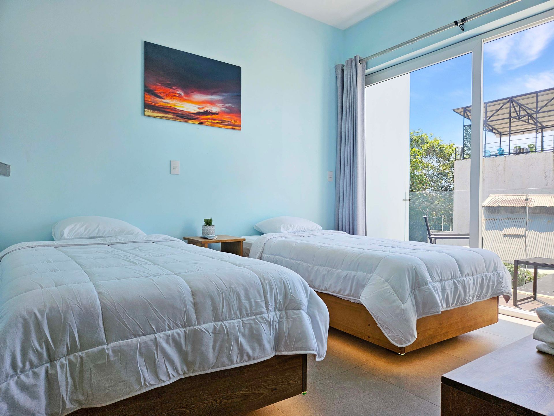 Two beds in a room with blue walls, a sunset painting, and a balcony view.