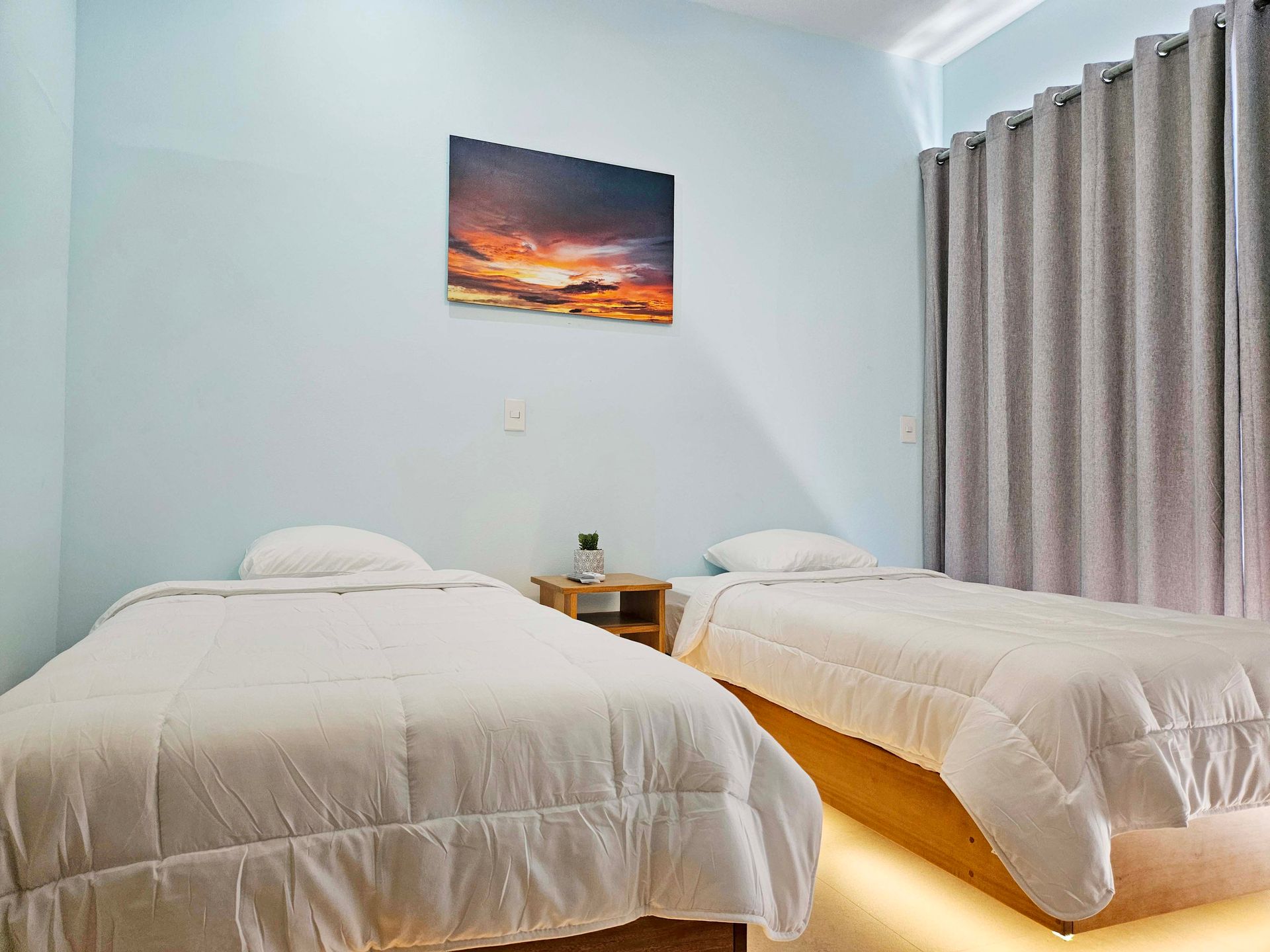 Two twin beds with white bedding in a room with blue walls, a sunset painting, and gray curtains.