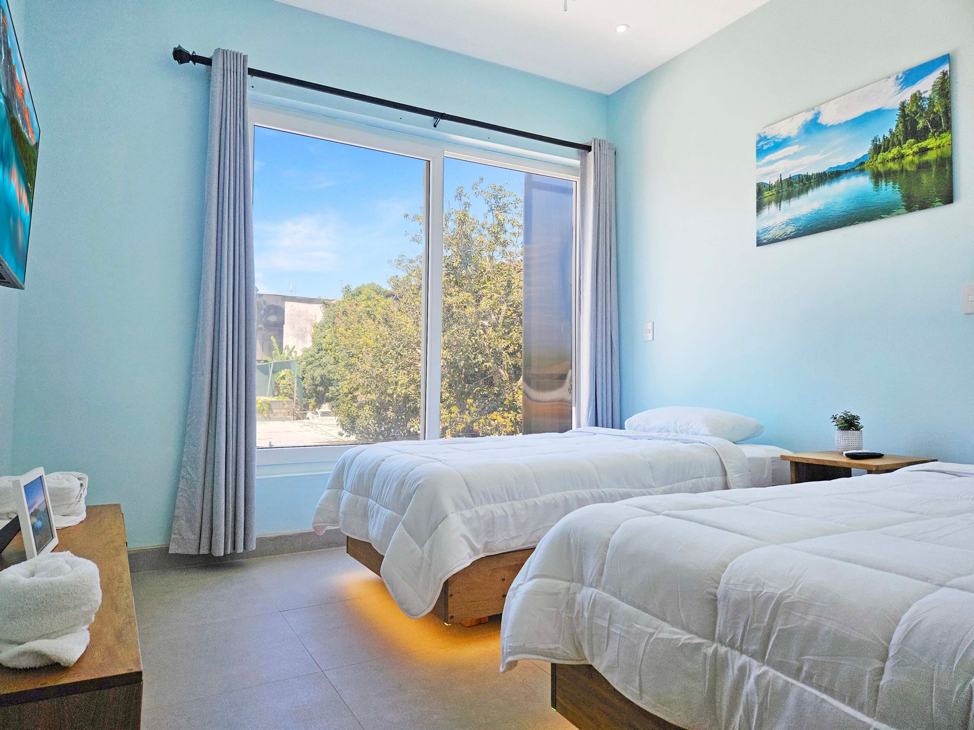 Bedroom with two twin beds, large window, light blue walls, art, and a nightstand.