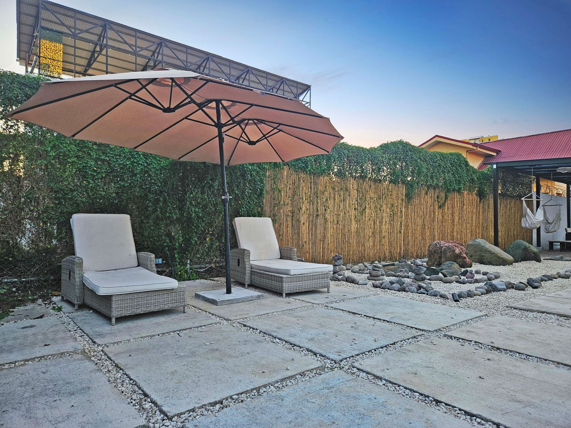 Two lounge chairs under an umbrella on a stone patio, with a bamboo and hedge backdrop.