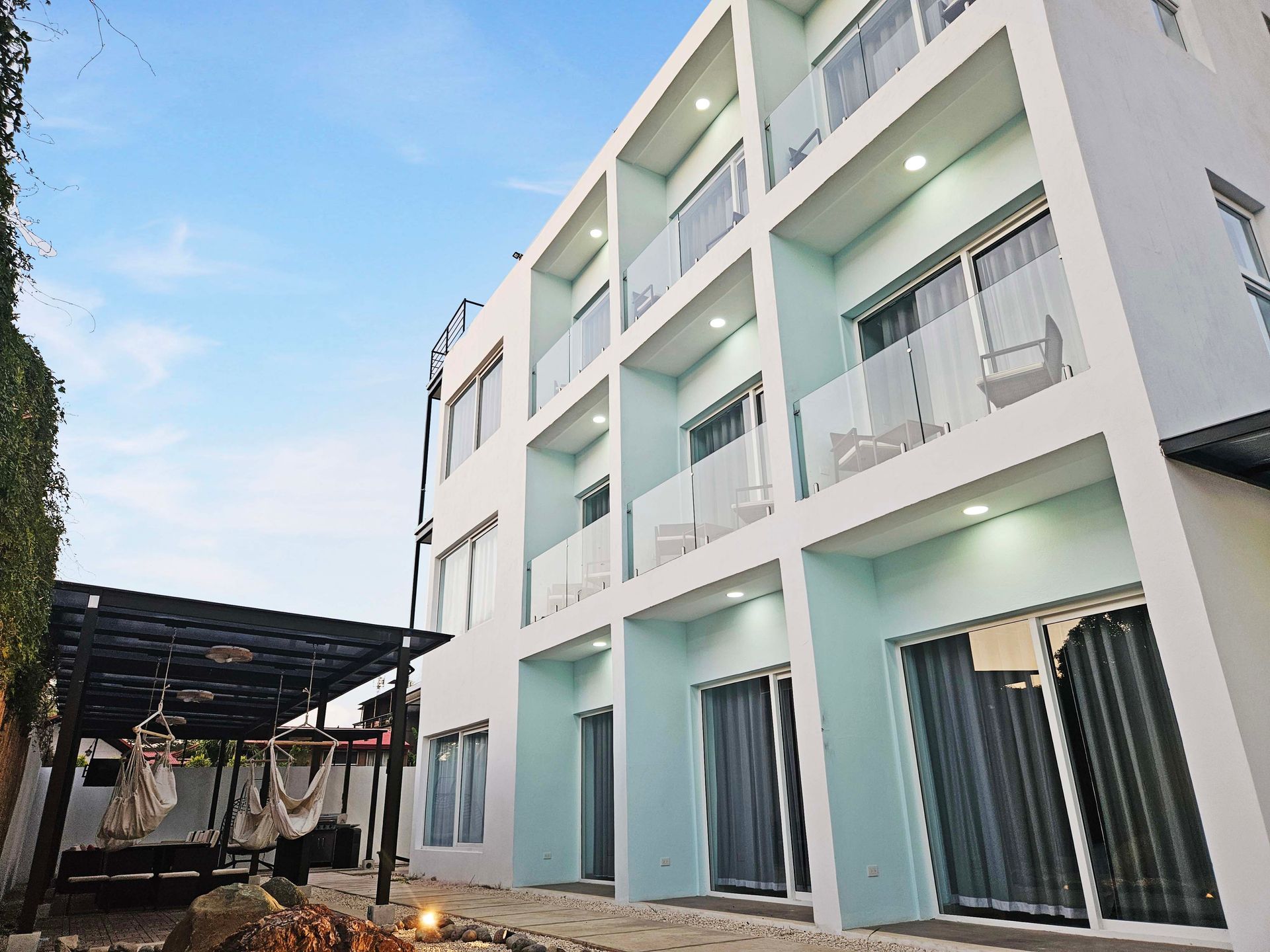 Light blue multi-story building with glass balconies, outdoor seating, and a shaded pergola.
