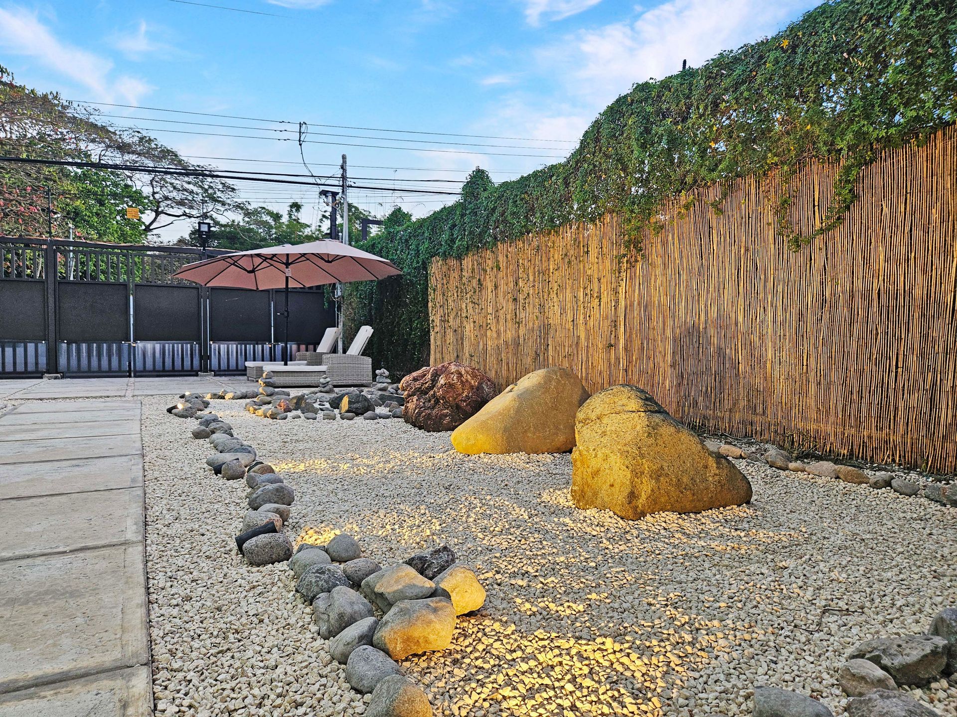 Zen garden with beige gravel, large rocks, bamboo fence, and lounge chairs under an umbrella.