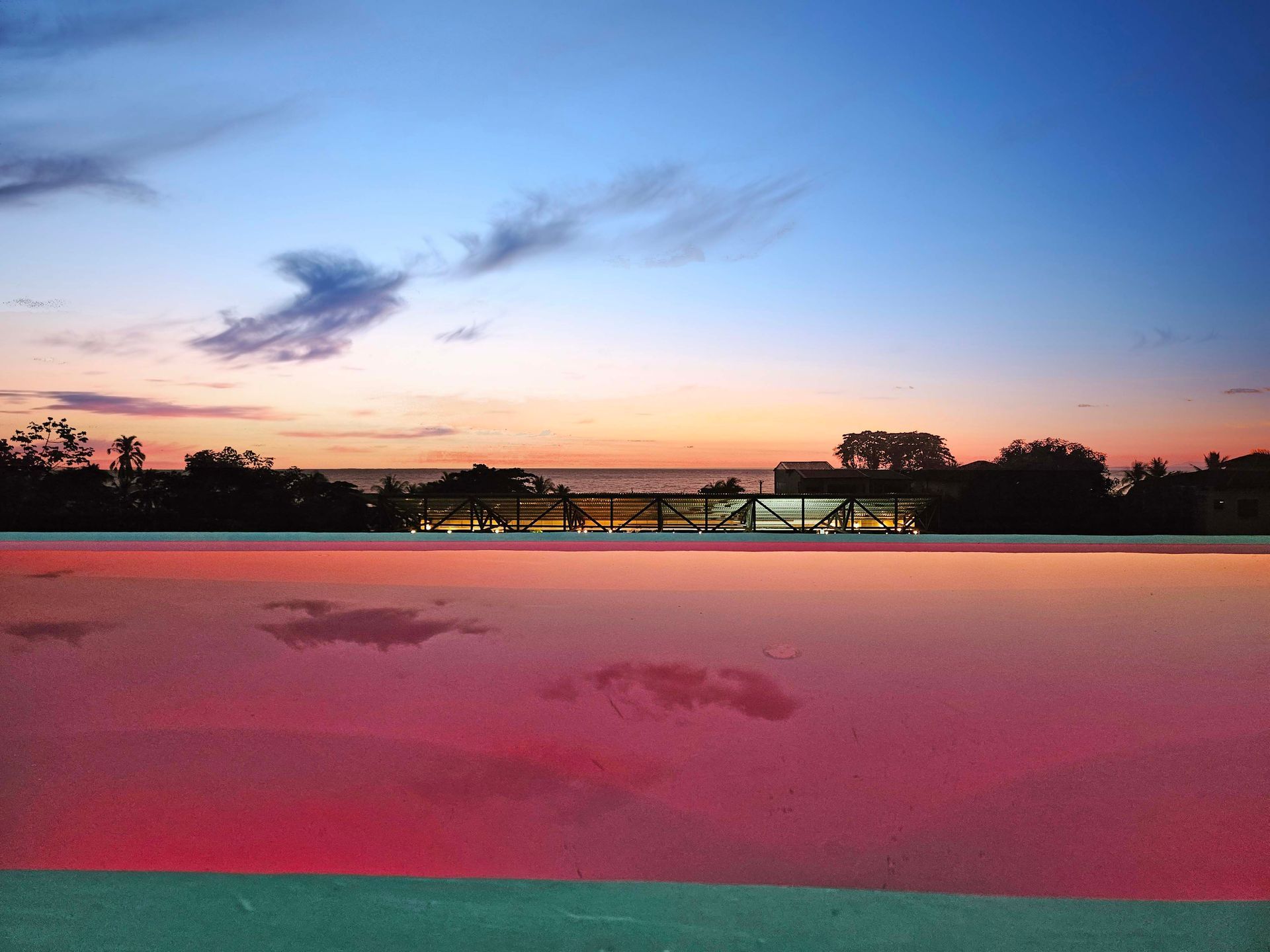 Sunset sky reflected in a red-tinted pool. Silhouetted trees and buildings along the horizon.