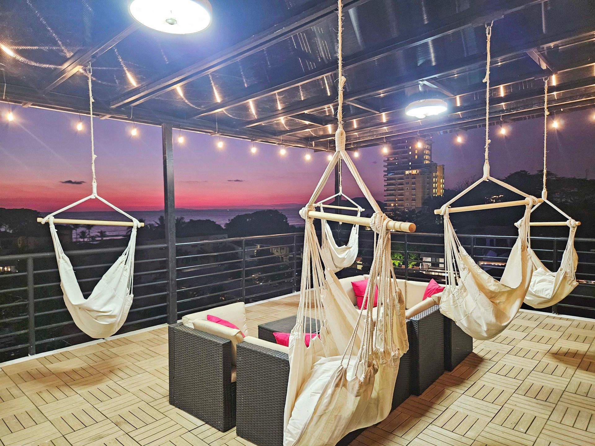 Rooftop lounge with hanging chairs, sofa, and city view at sunset.
