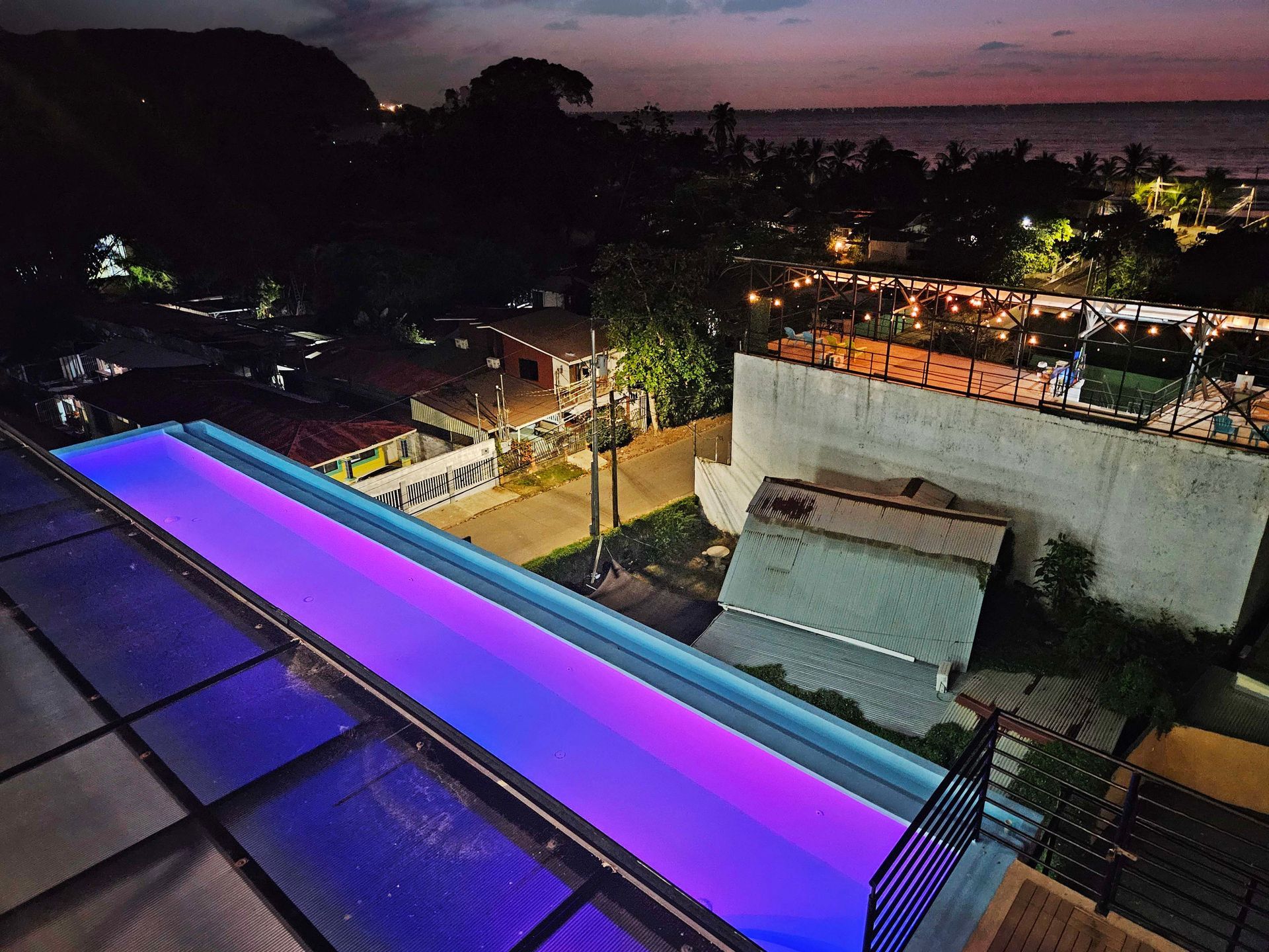 Rooftop pool glowing purple at dusk, overlooking city buildings and ocean.