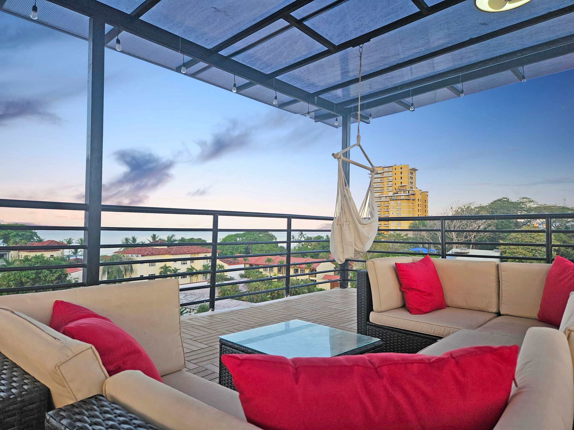 Rooftop patio with seating, red pillows, glass table, hammock, and city view at dusk.