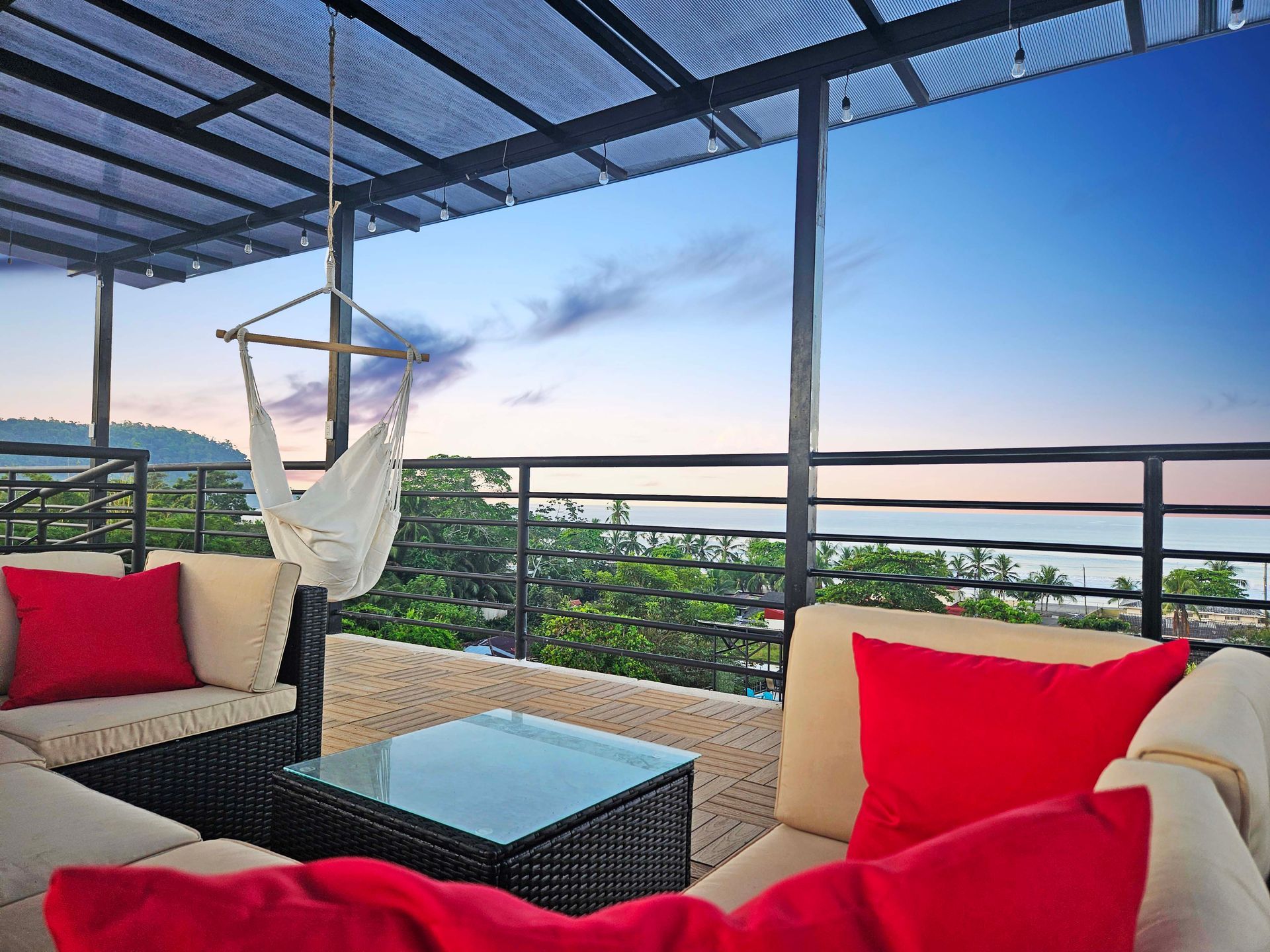 Rooftop patio with seating, hammock, and ocean view at sunset. Red pillows and blue sky.