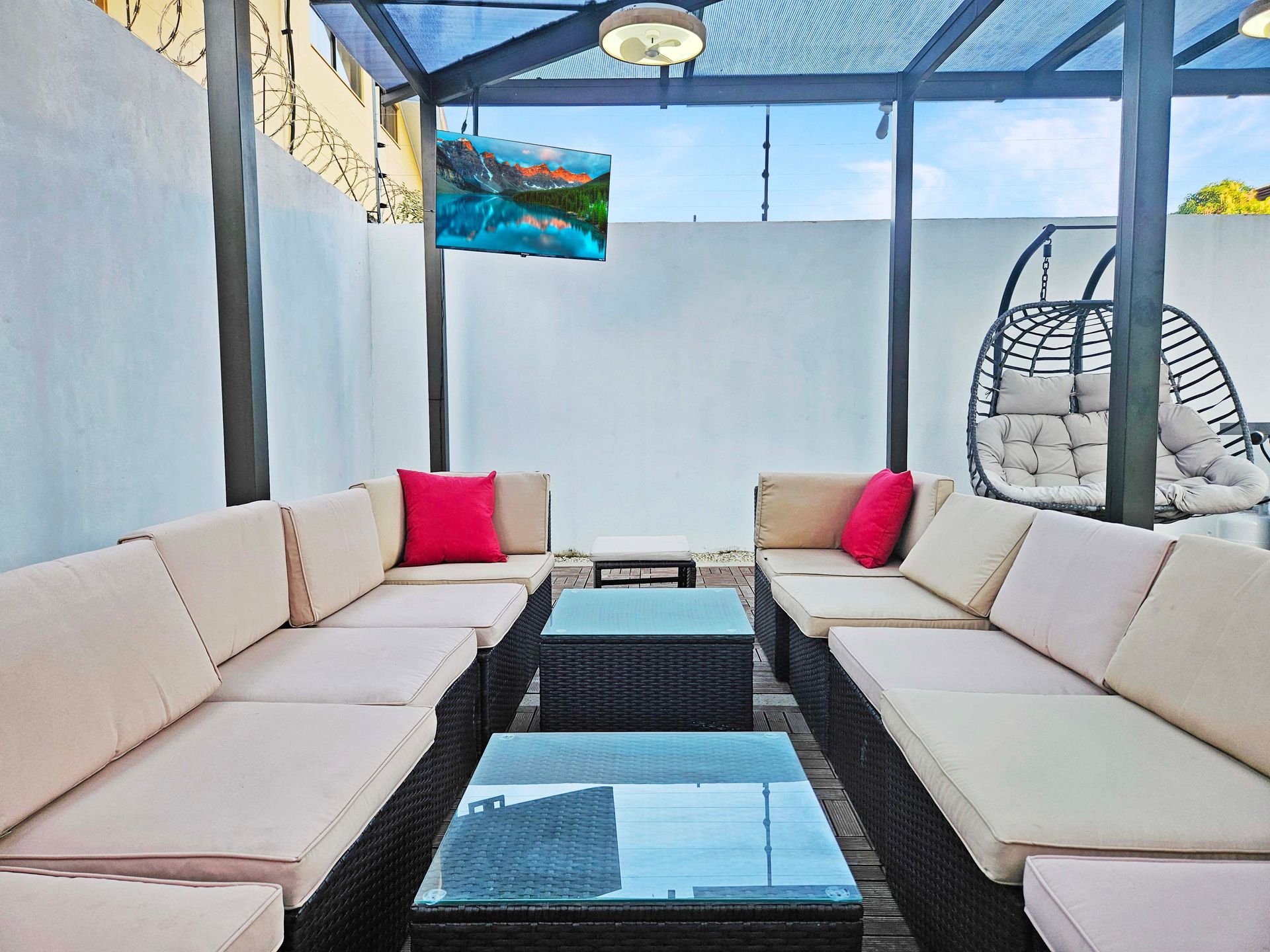 Outdoor lounge area with beige sofas, red pillows, glass tables, a hanging chair, and a TV.