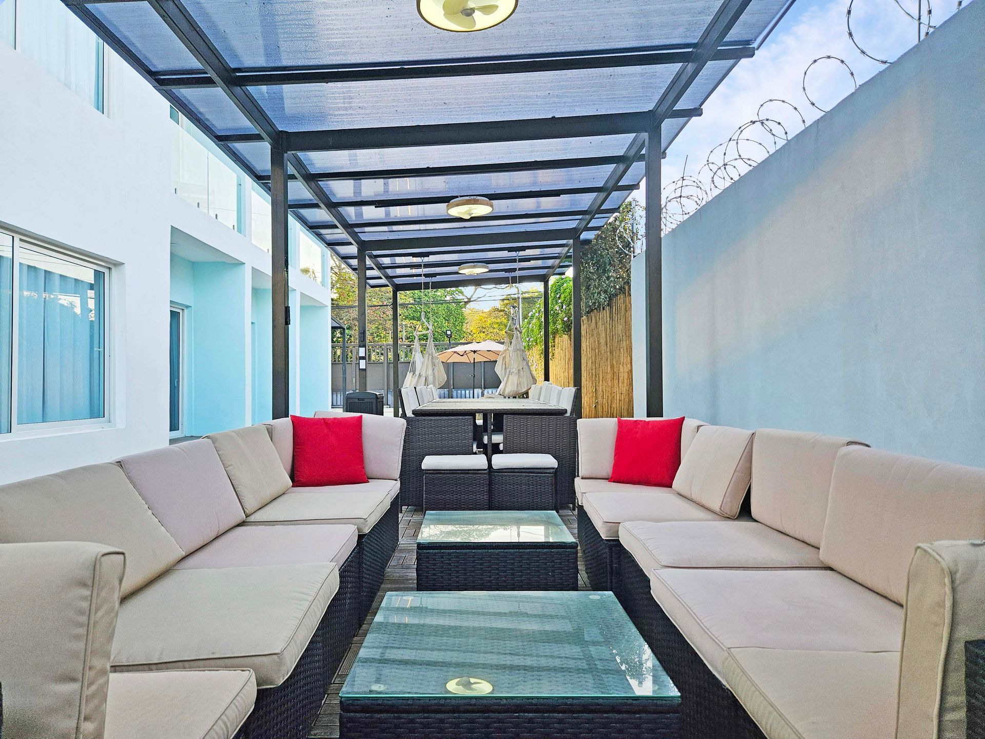 Outdoor seating area with beige couches, red pillows, and glass coffee tables under a covered walkway.