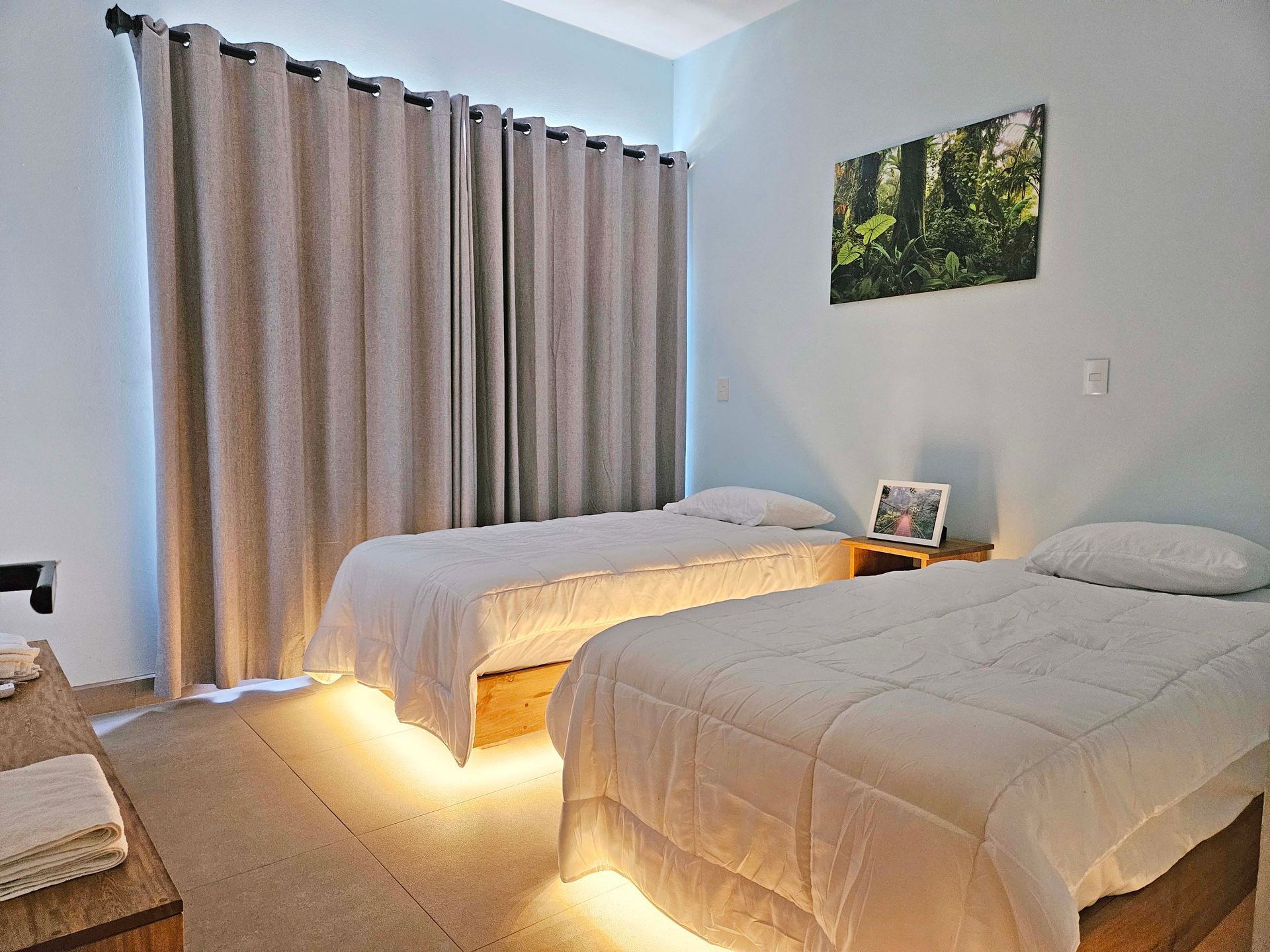 Two twin beds with white bedding in a light blue room, lit by under-bed lights, with curtains and artwork.