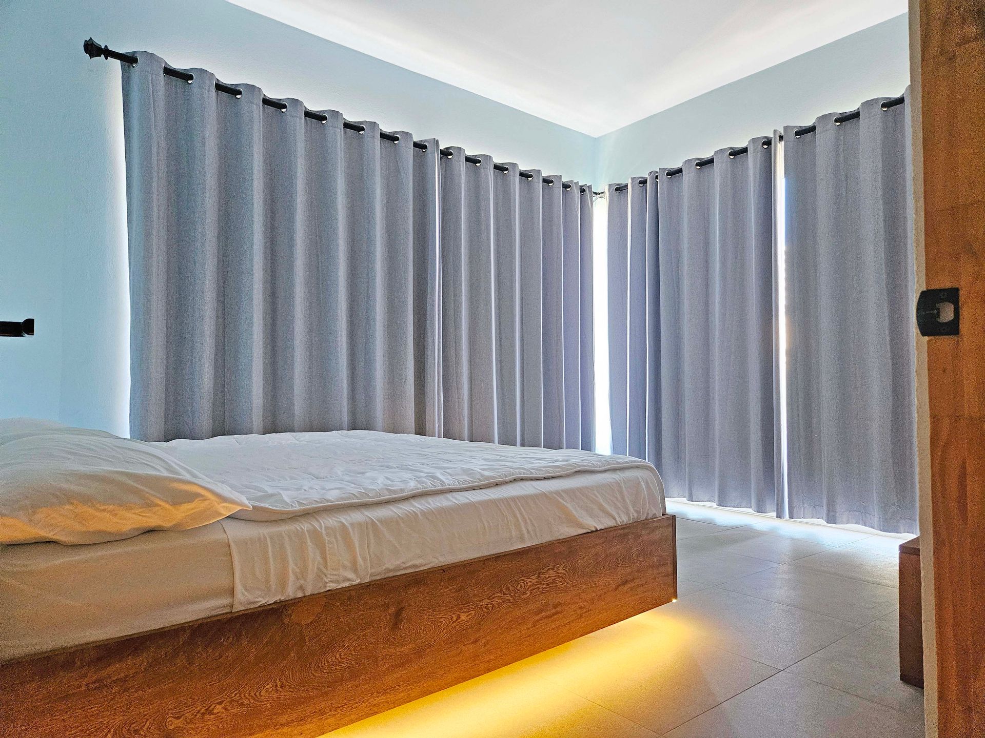Bedroom with light blue walls, gray curtains, a wooden bed frame with under-bed lighting.