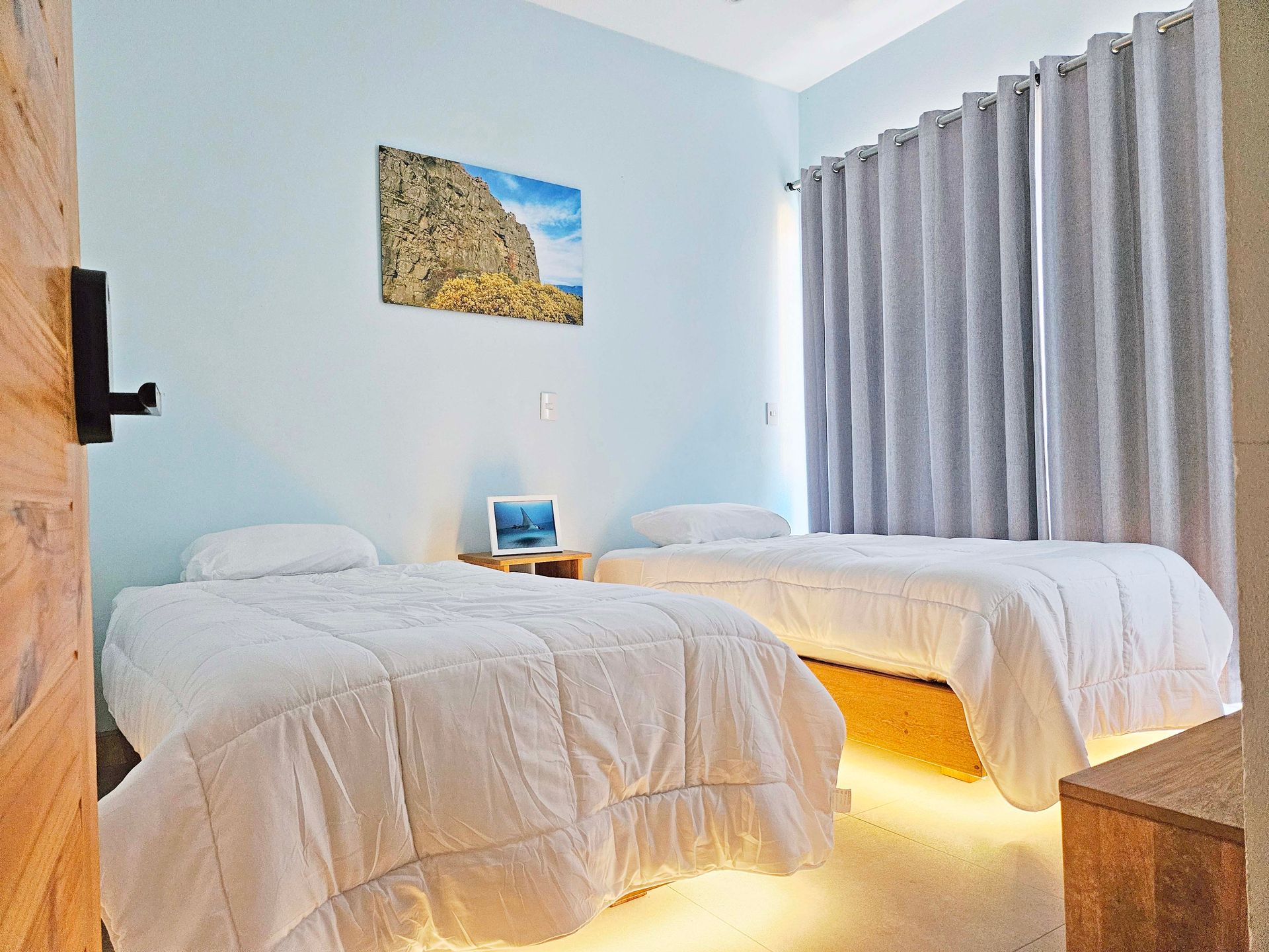 Twin beds in a light blue room with a landscape picture and light gray curtains.