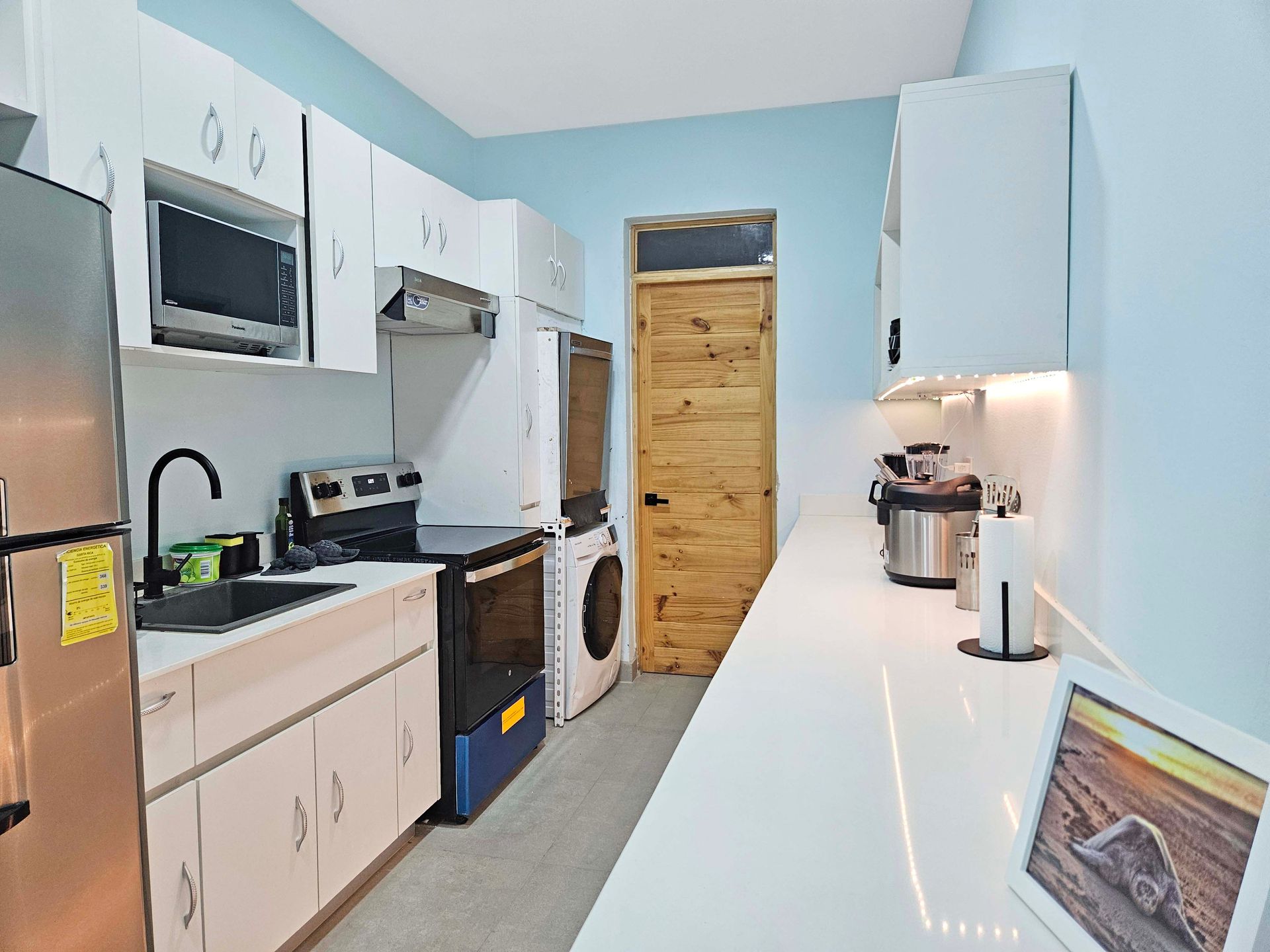 Small, white kitchen with appliances and white countertops, blue walls, wooden door.