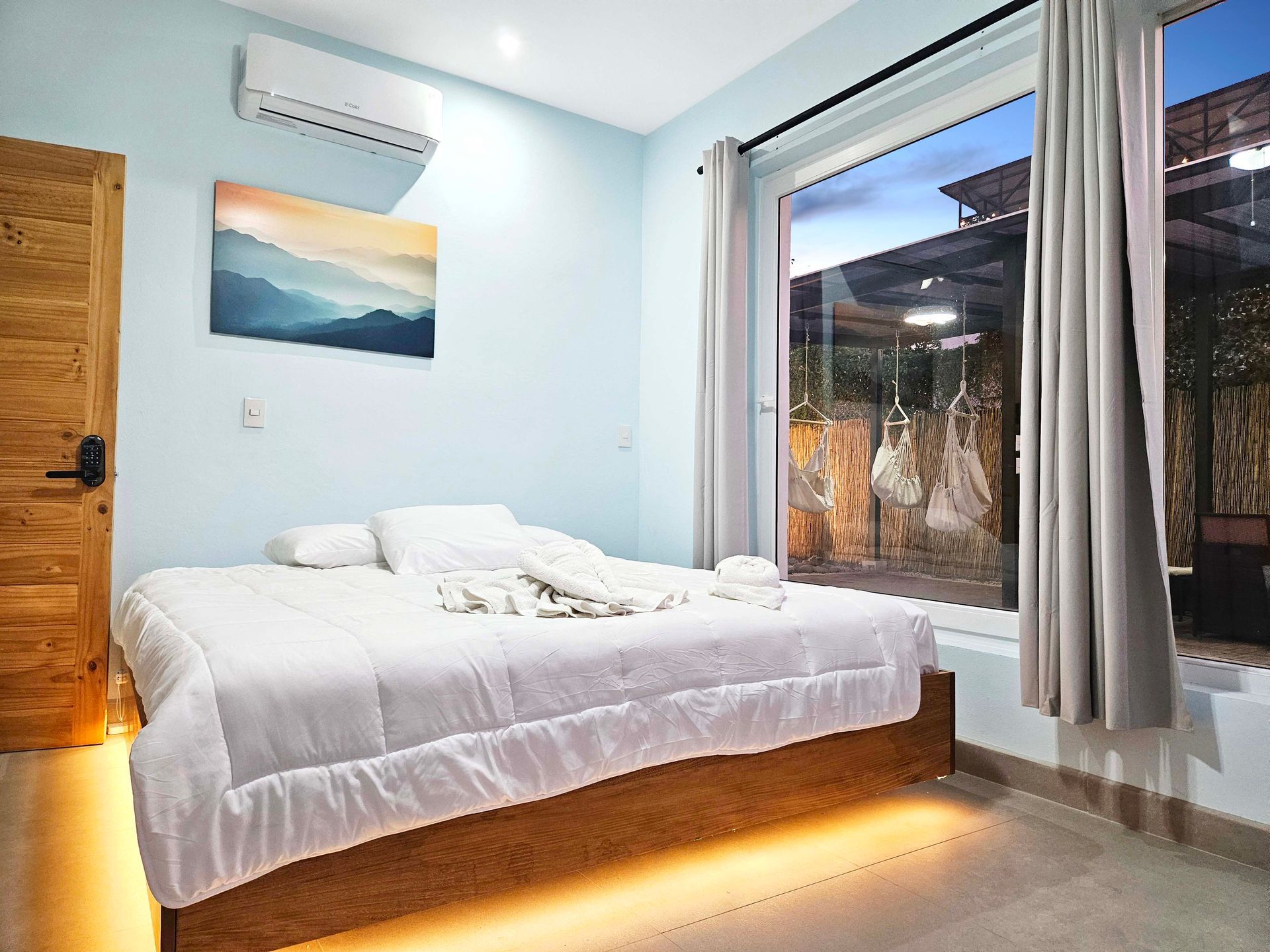 Bedroom with a white bed, wooden door, and a window overlooking an outdoor area at dusk.