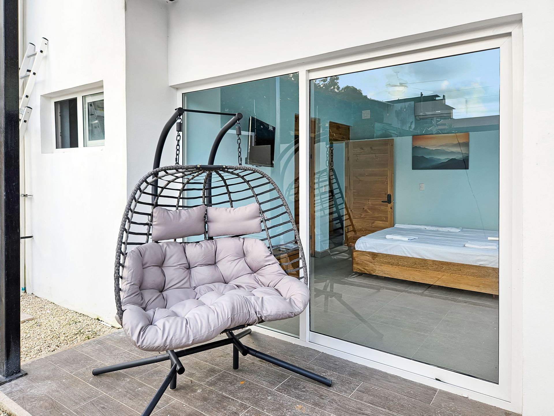 Wicker hanging chair on patio, sliding glass door reveals bedroom with bed and artwork.