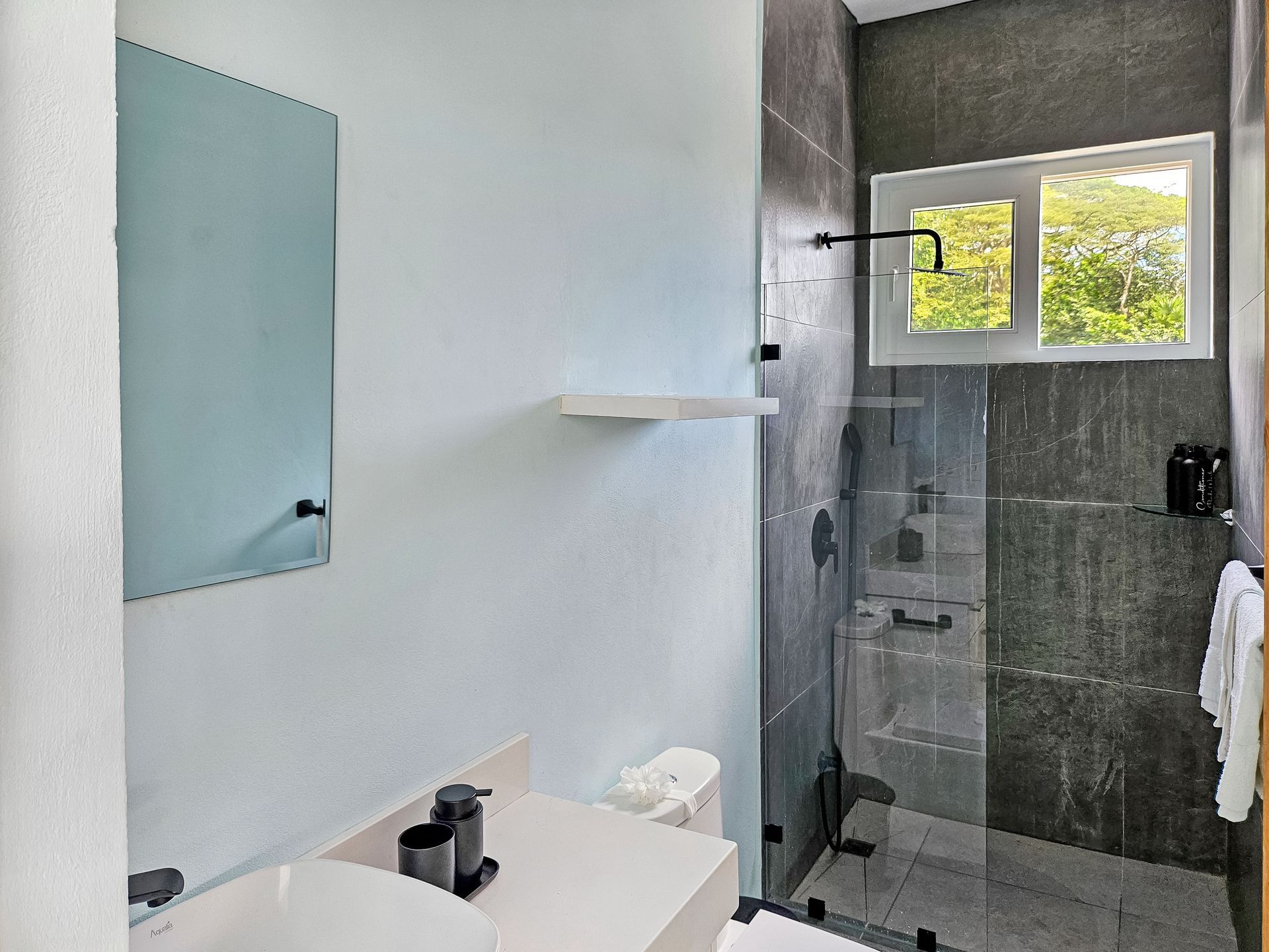 Modern bathroom with gray tiled shower, window, white sink, and light blue wall.