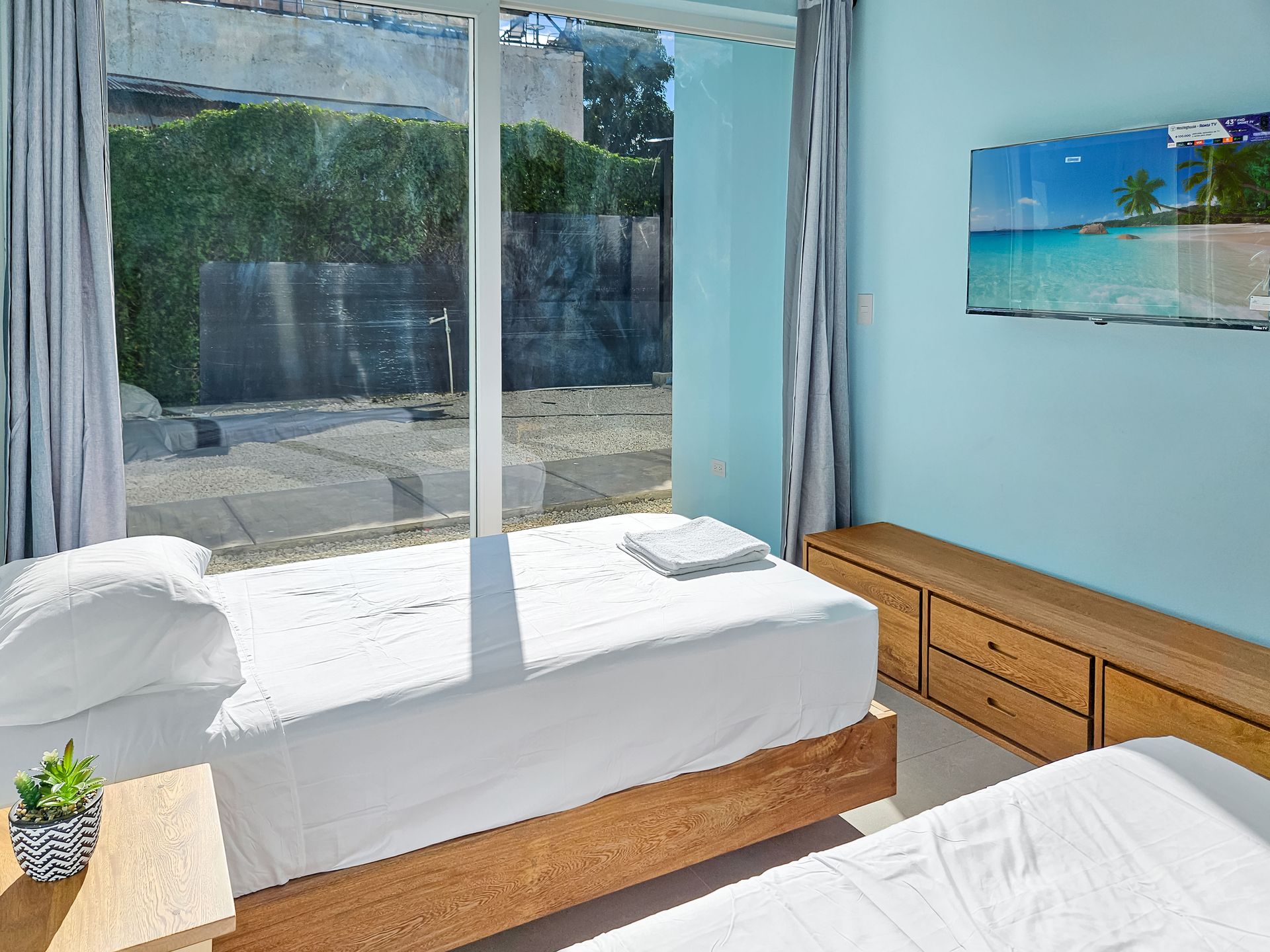 Twin beds in a light blue bedroom, large window with outdoor view, wood bedside table, beach scene art.
