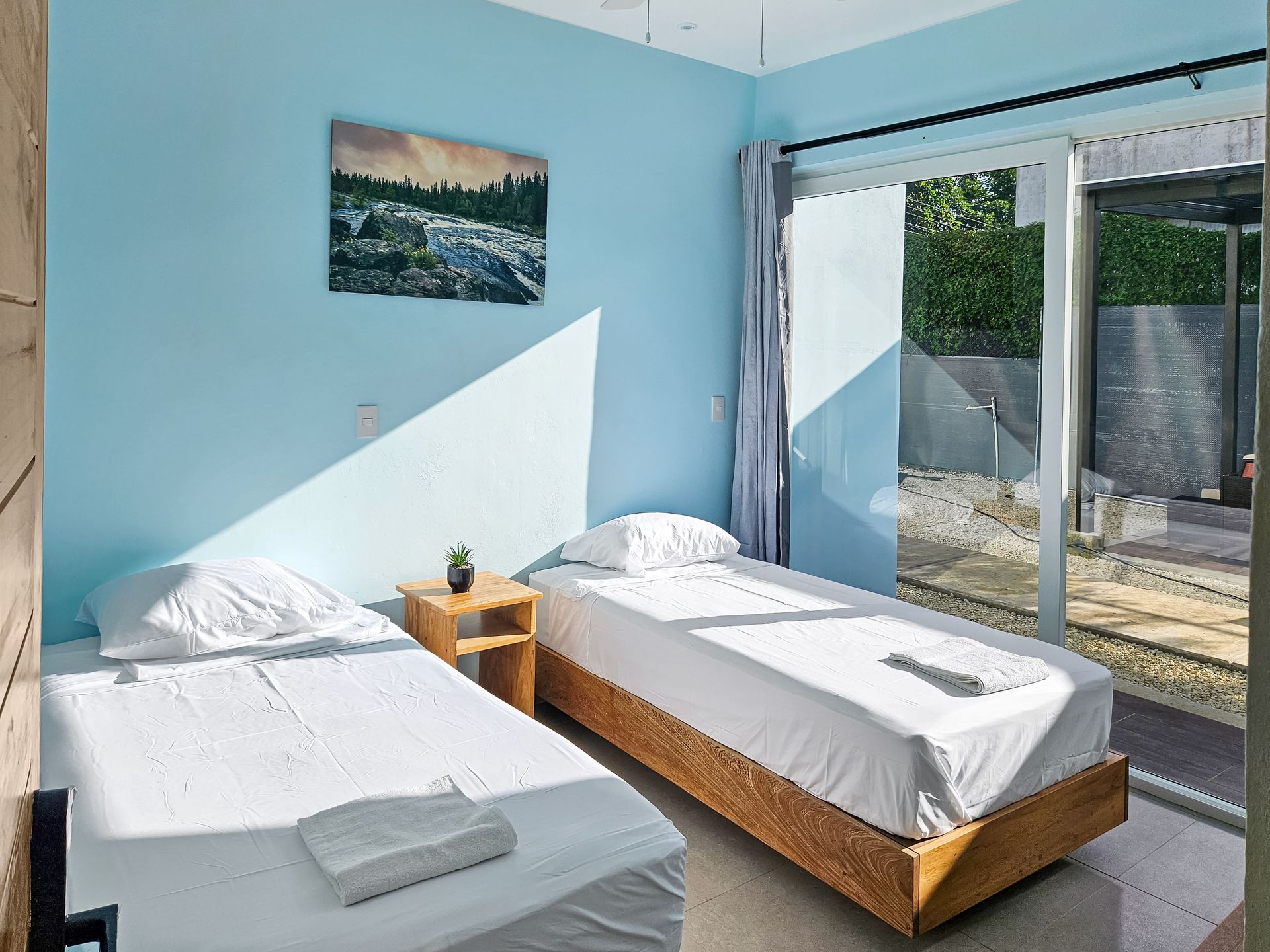 Two twin beds in a room with blue walls, sliding glass door, and wooden nightstand with a painting.