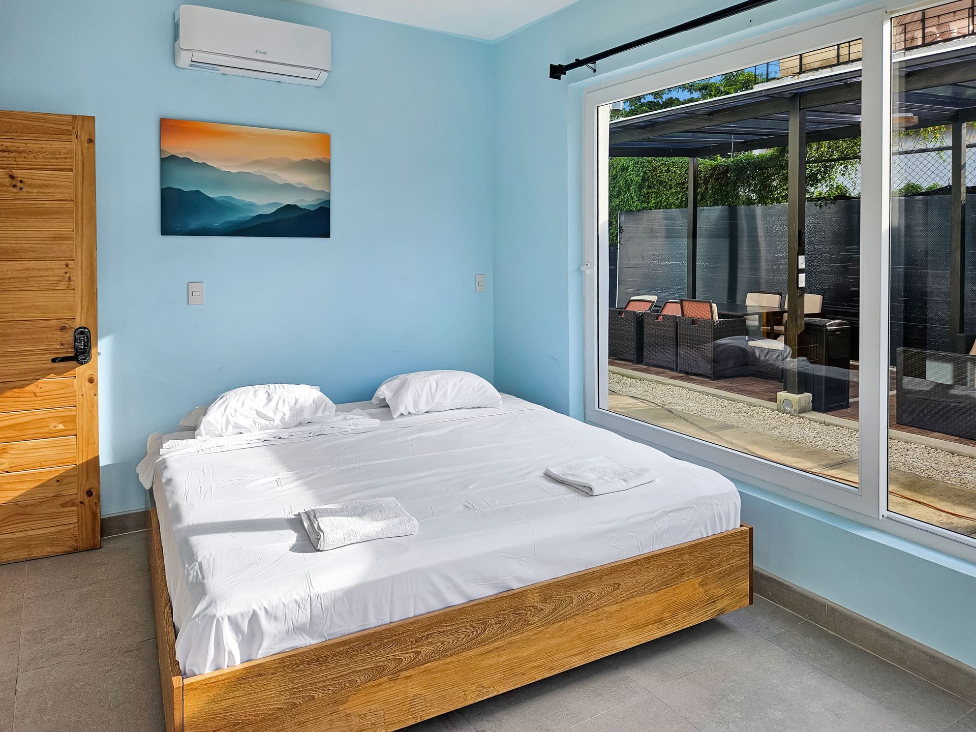 Bedroom with a wooden bed, white linens, and a view of an outdoor patio through a large window.