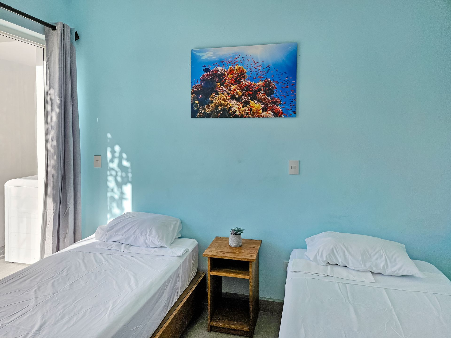 Two twin beds in a room with light blue walls and a coral reef painting. A small wooden nightstand sits between the beds.