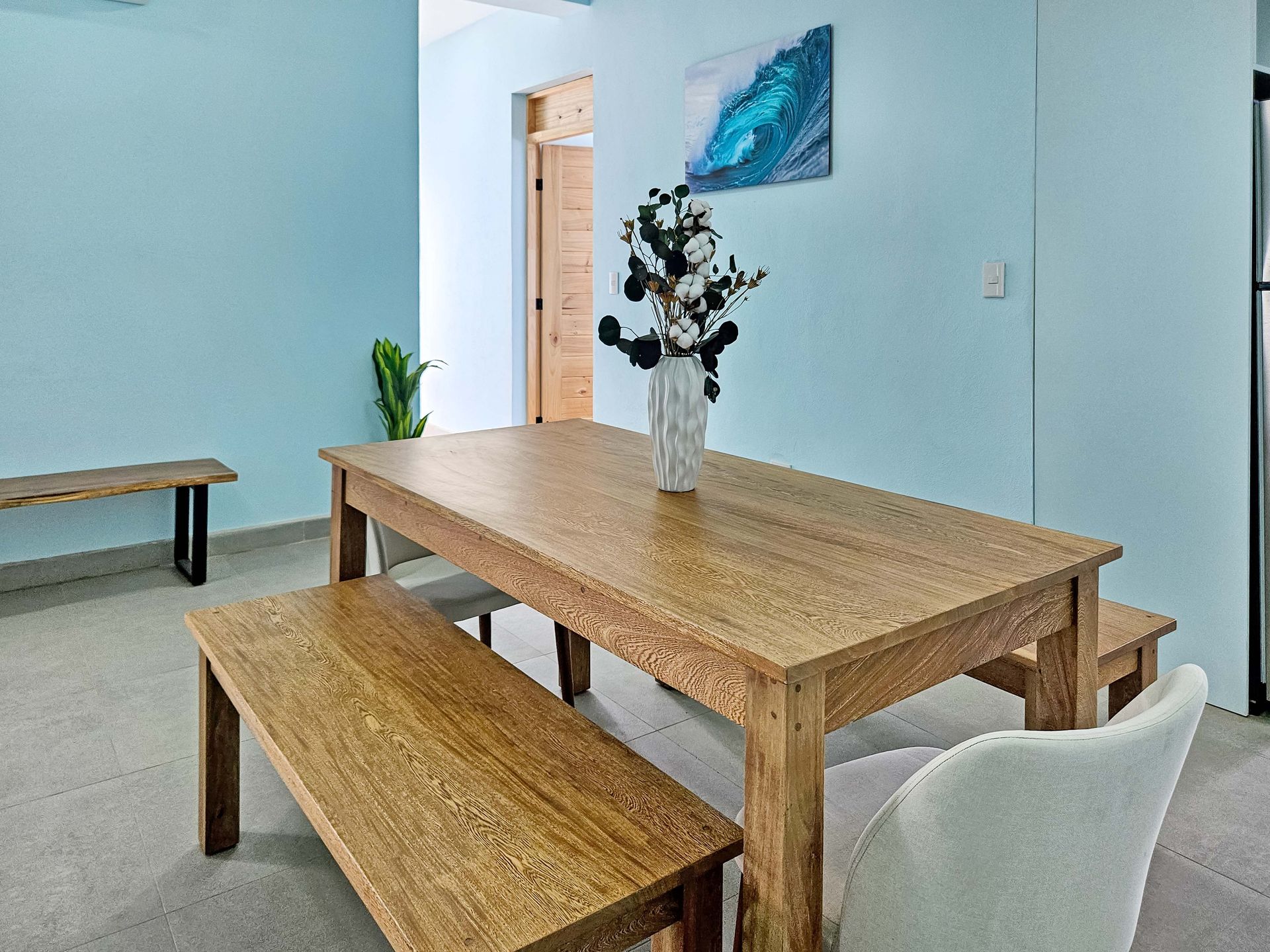 Dining room with wooden table and benches, light blue walls, art, and a vase of flowers.