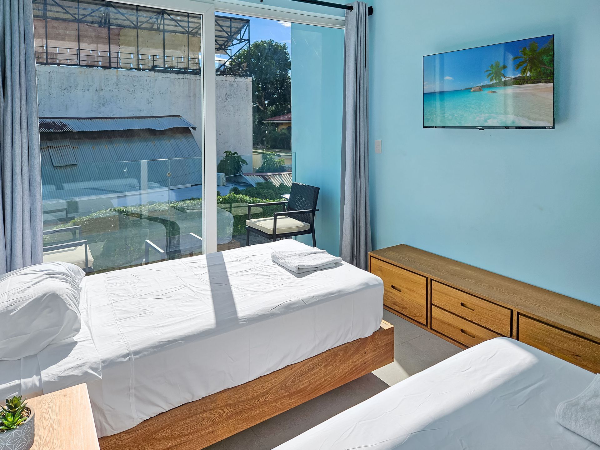 Bedroom with two beds, large window, TV, light blue walls, and wooden furniture.