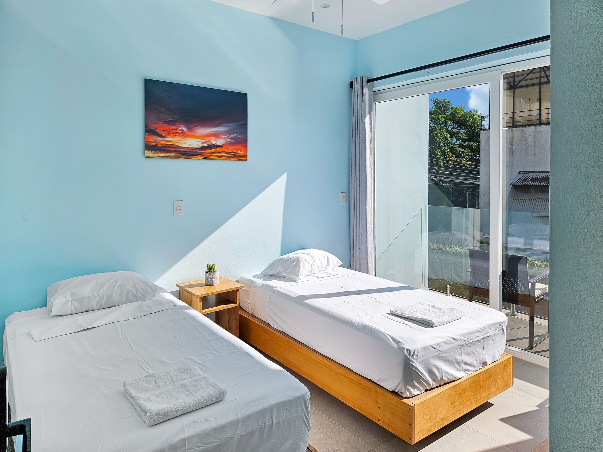 Two twin beds in a room with blue walls, a sunset painting, and a balcony.