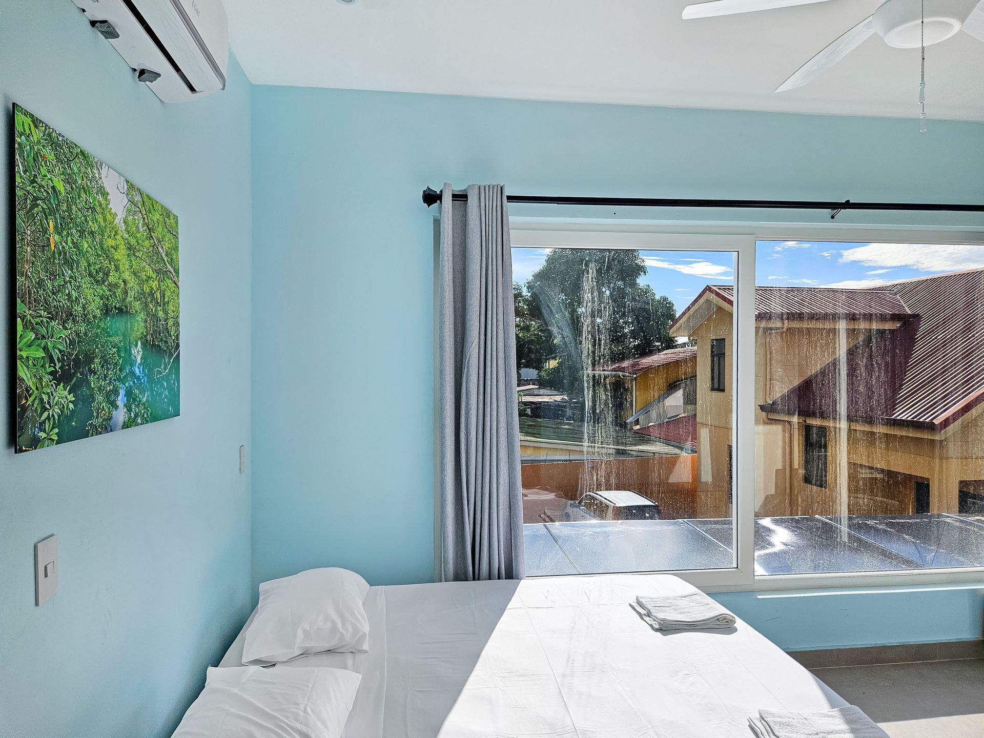 Bedroom with bed, blue walls, a large window with houses outside, and a painting.