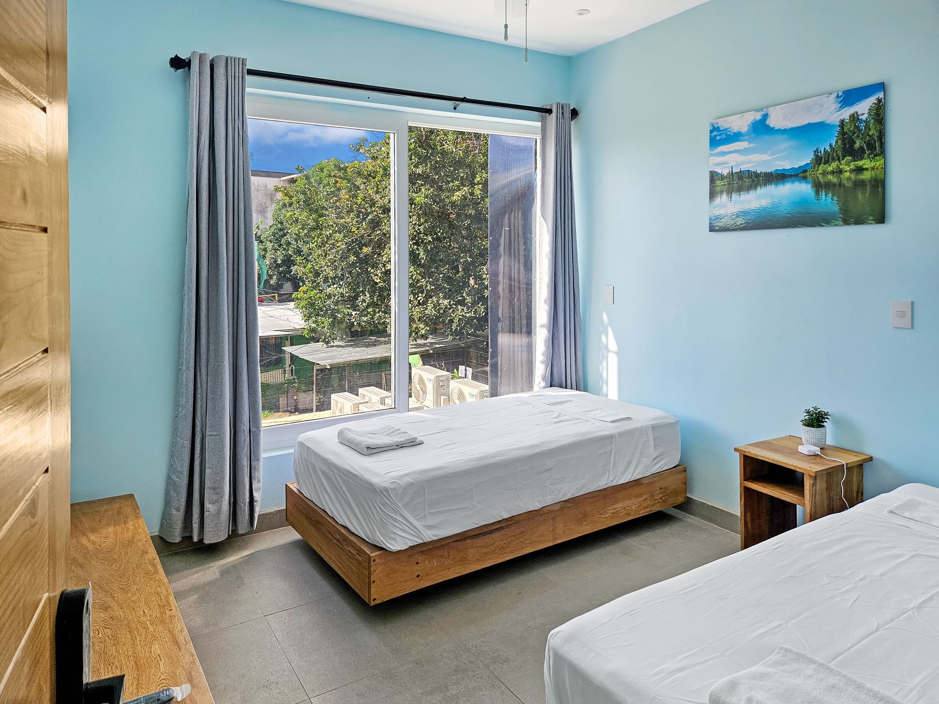 Bedroom with two twin beds, large window, light blue walls, art, and wooden nightstand.
