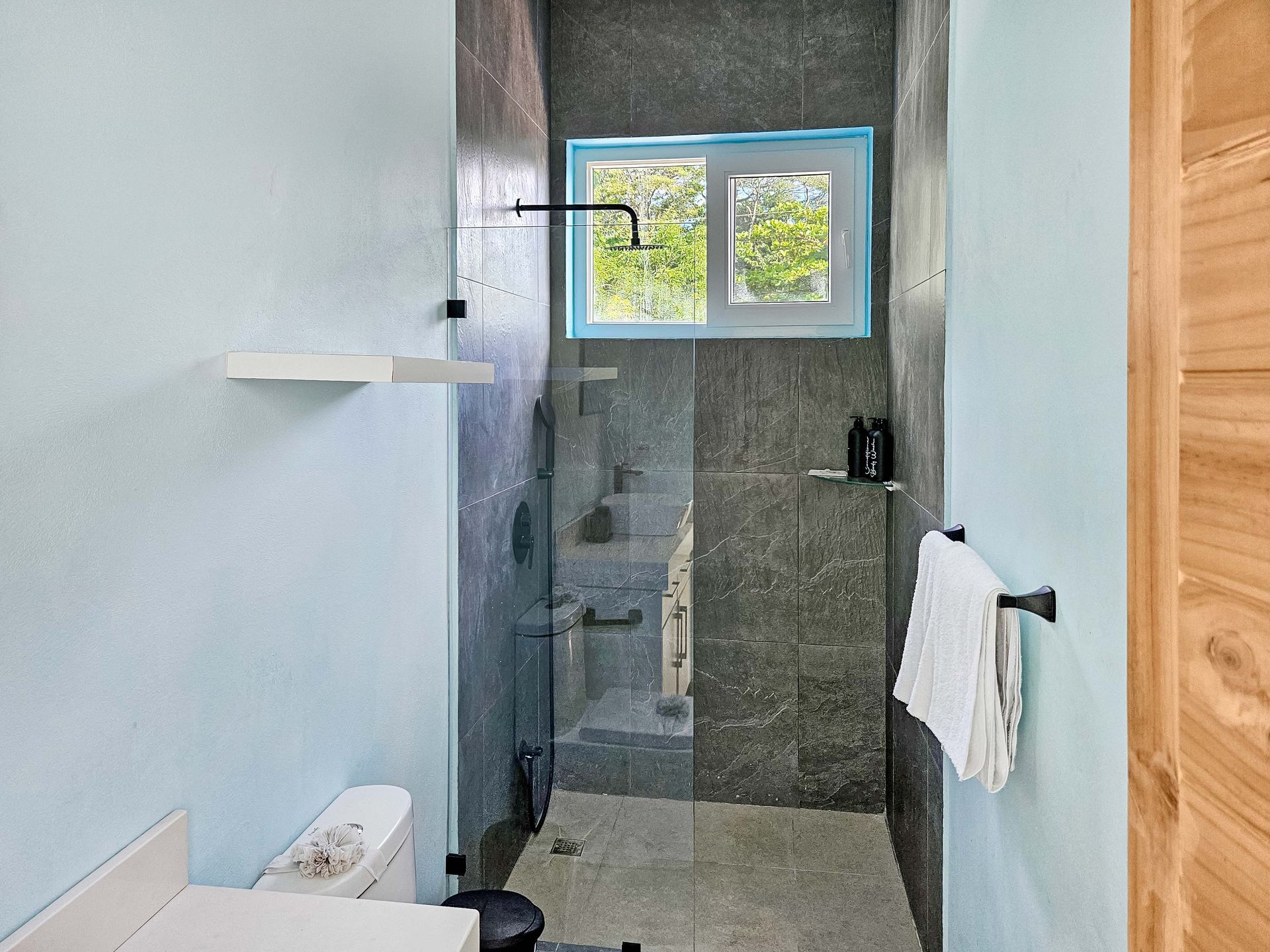 Modern bathroom with a glass-enclosed shower, dark gray tile, window, toilet, and light blue walls.