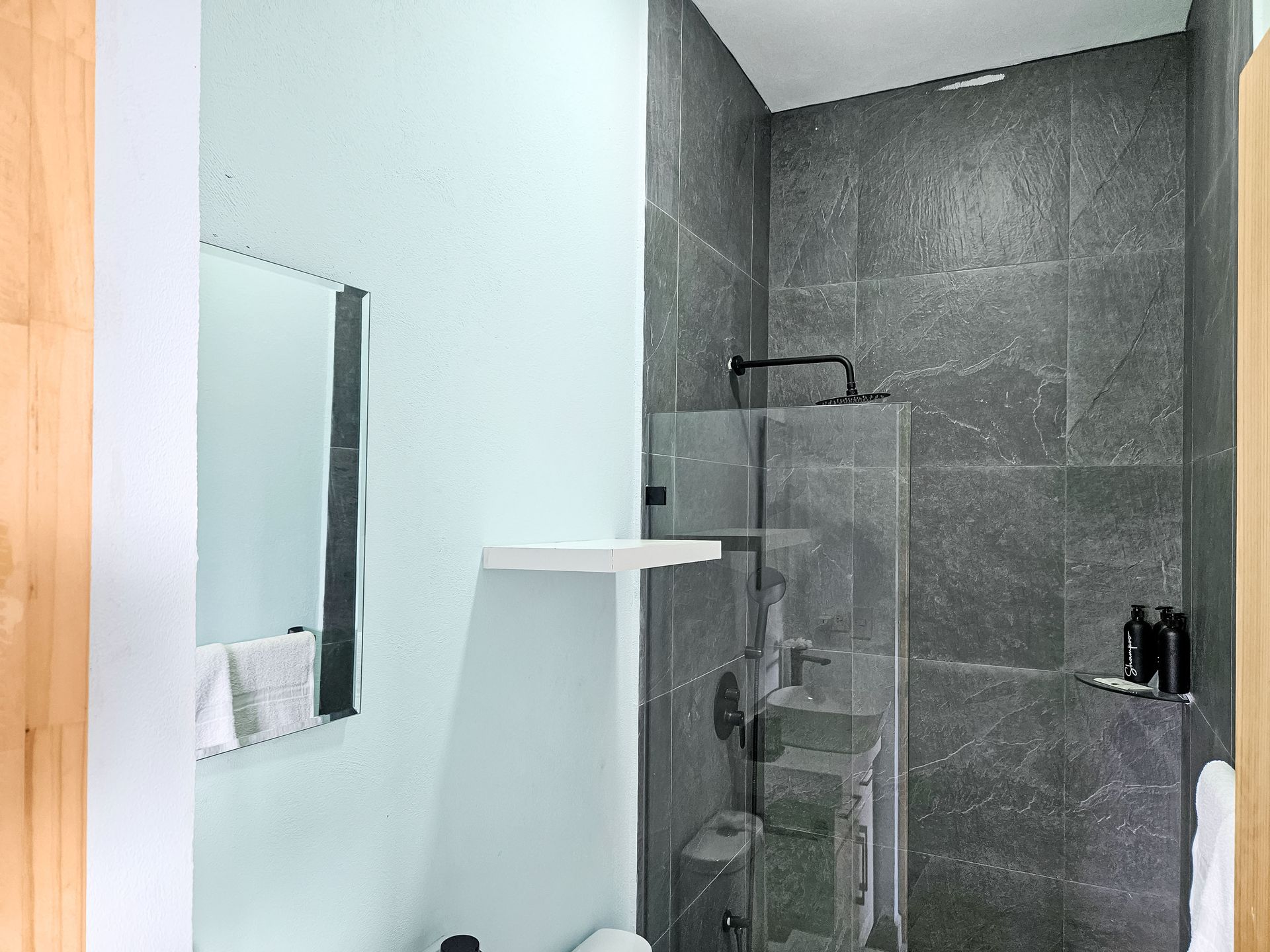 Bathroom with gray tile shower and glass door, pale blue wall, mirror.