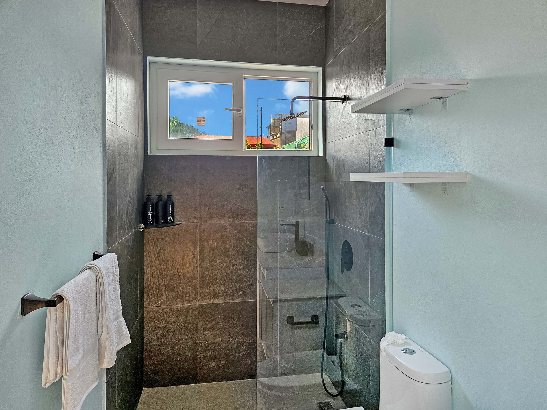 Small bathroom with a shower, toilet, and window showing a sunny view. Gray tile and pale blue walls.